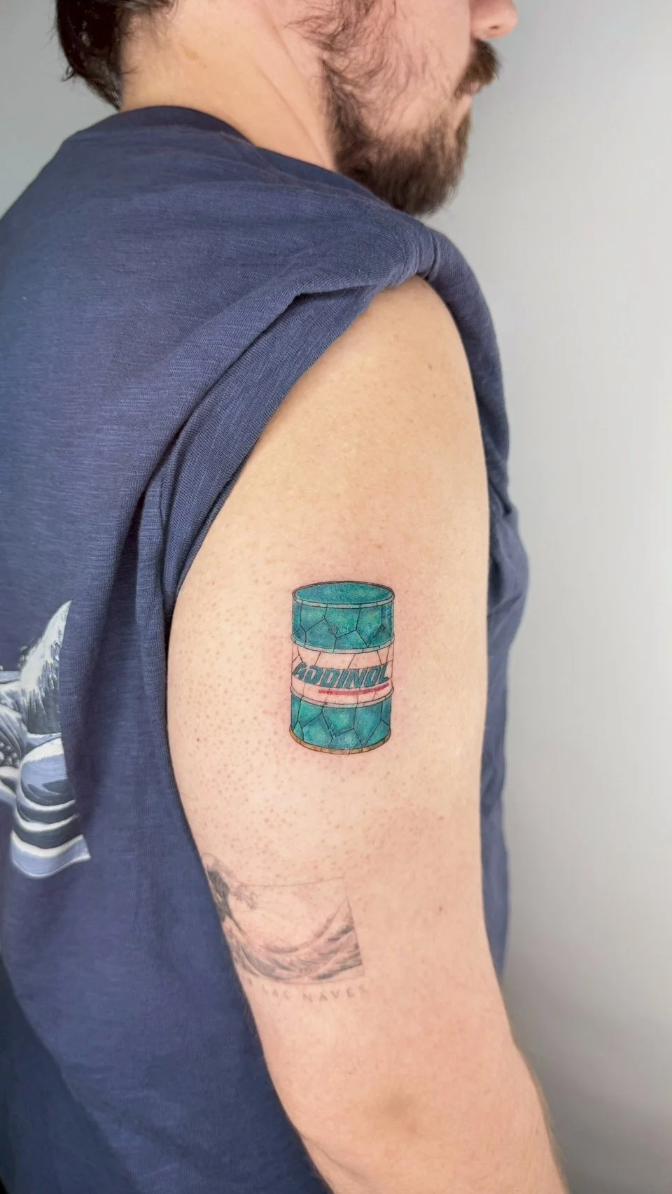 Tattoo of a turquoise, cracked soda can labeled 'ADDINOL' on the upper arm of a man with a beard, wearing a sleeveless shirt.