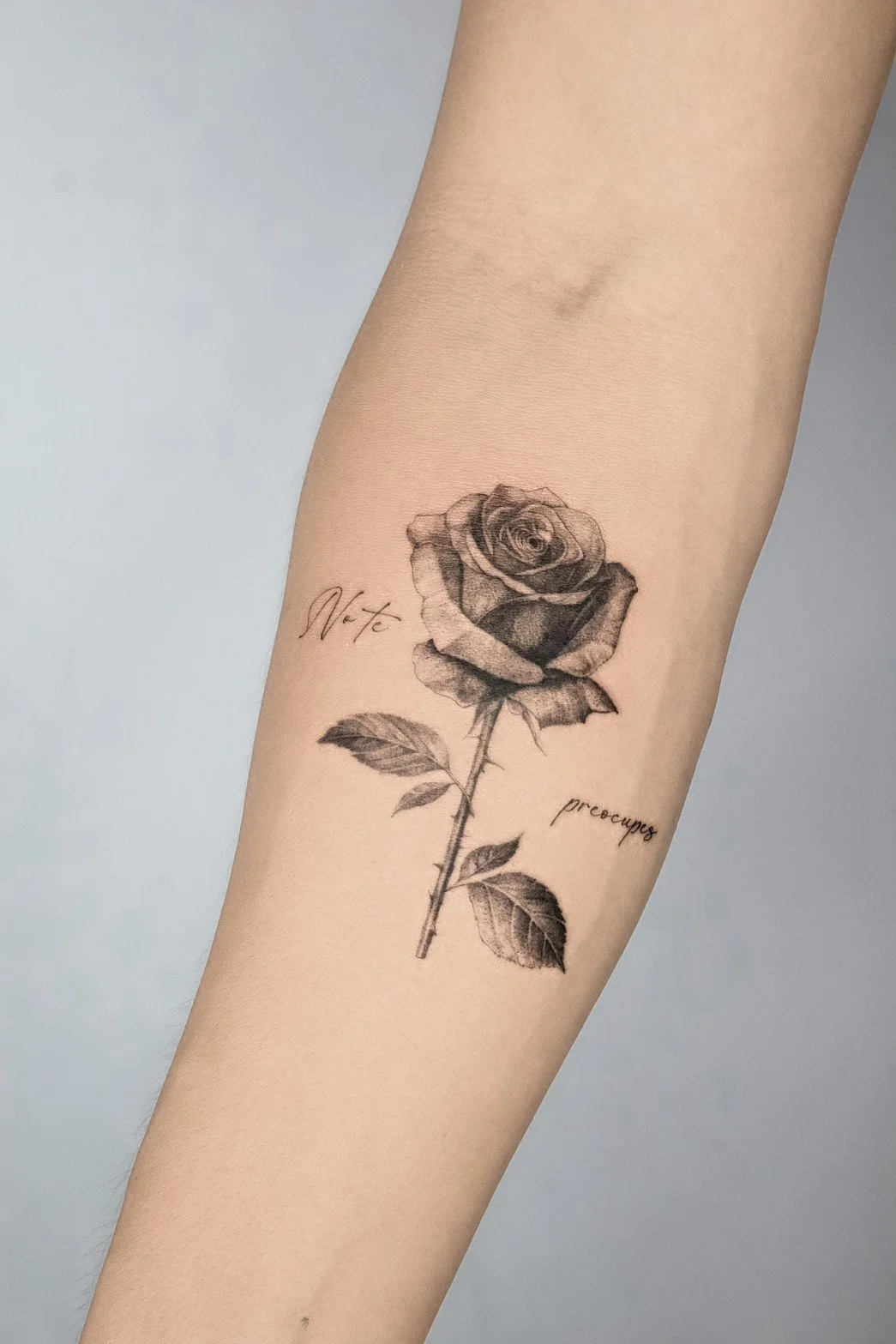 Tattoo of a realistic black and gray rose with leaves on forearm, with words 'Note' and 'preoccupy' written near it.