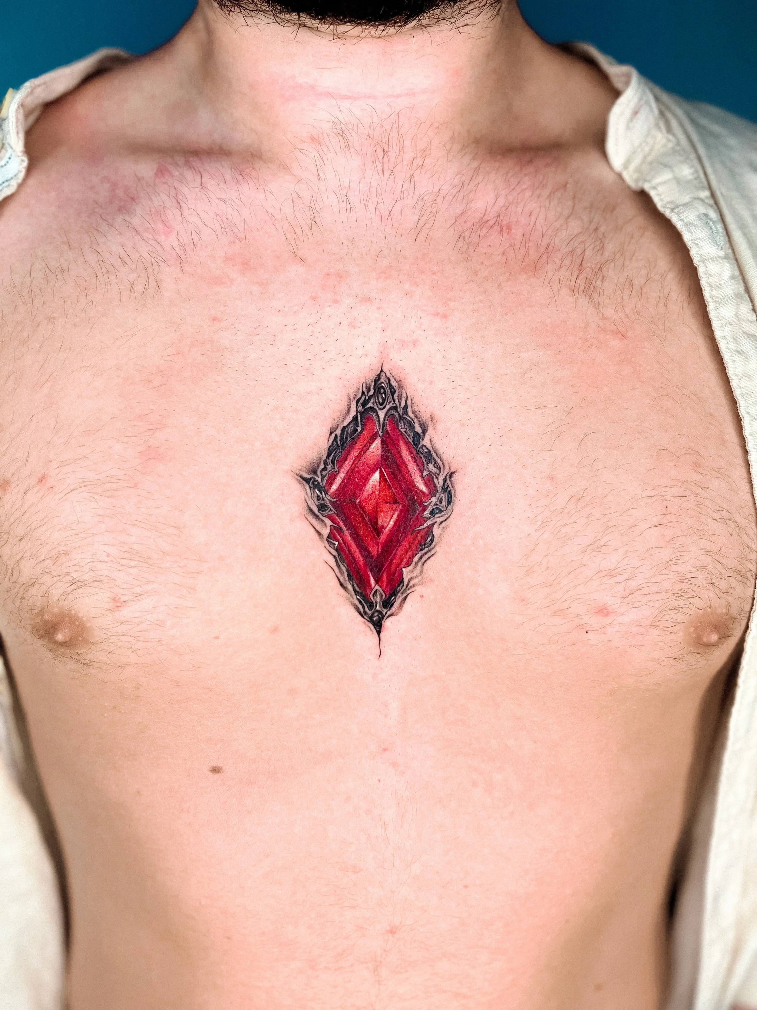 Close-up of a person's bare chest with a large tattoo of a red diamond-shaped gemstone at the center, outlined in black, with shading and details. The person is shirtless, and part of a light-colored shirt is visible on the sides.