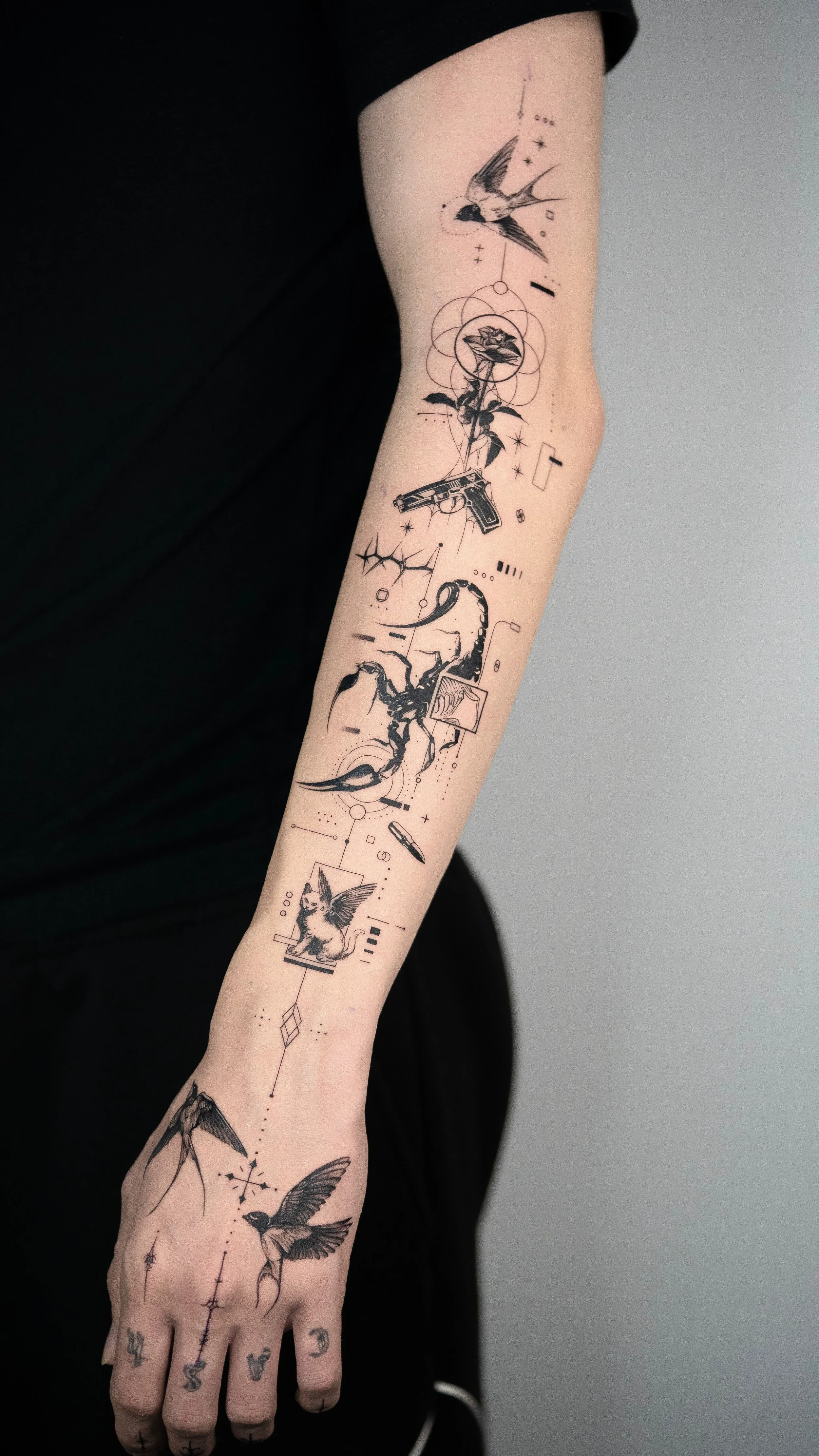 Black ink tattoo sleeve featuring a swallow, geometric shapes, a pistol, a serpent, a kitten with wings, and a hummingbird on a person's arm.