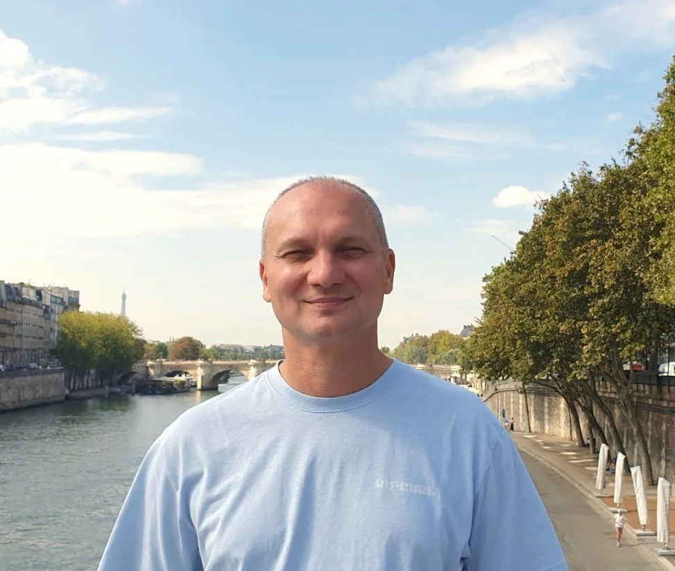A man with a shaved head smiling outdoors by a river, with bridges and buildings in the background under a partly cloudy sky.