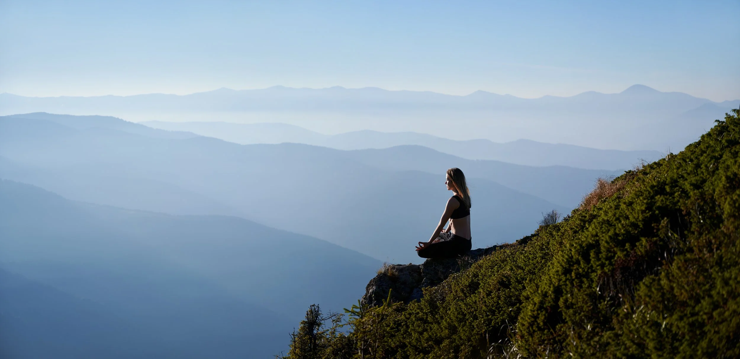 A woman practicing meditation on a rocky outcrop overlooking a series of layered blue mountains under a clear sky.