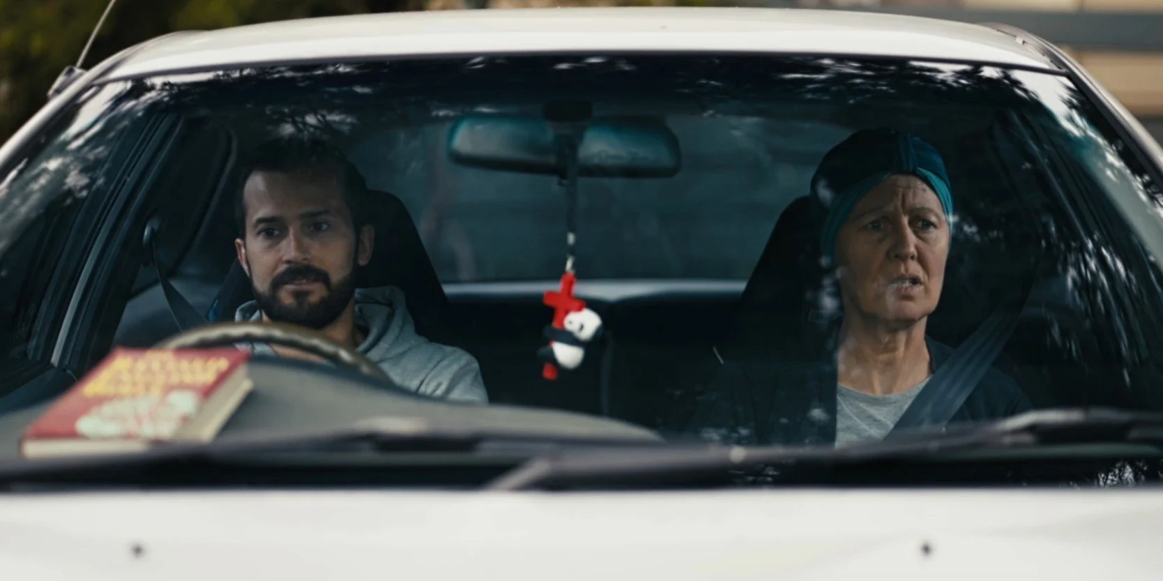 Two people sitting in a car viewed from the front windshield. A man is driving, and a woman is in the passenger seat. A red and black air freshener hangs from the rearview mirror, and there is a book on the dashboard.