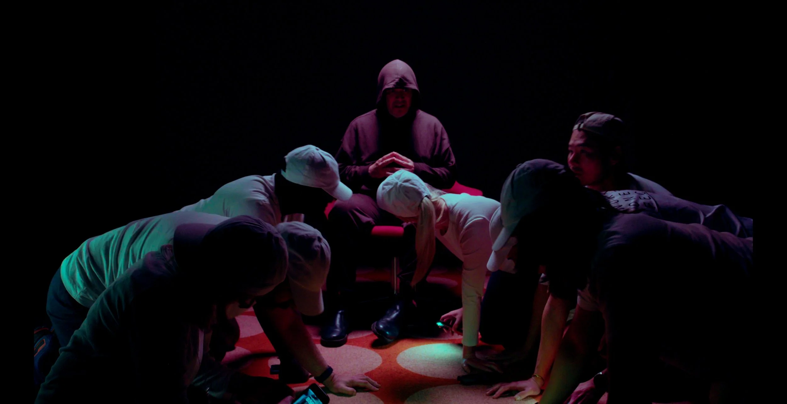 People kneeling in front of a hooded figure on a chair in a dark room.