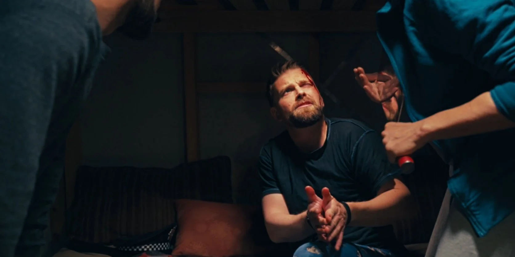 A tense scene with a man with a cut on his forehead, sitting between two others in a dimly lit room, one holding a screwdriver.