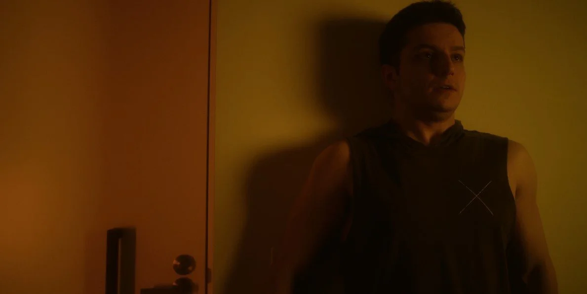 A person standing in dim lighting against a yellow wall, near a closed door with visible shadows.