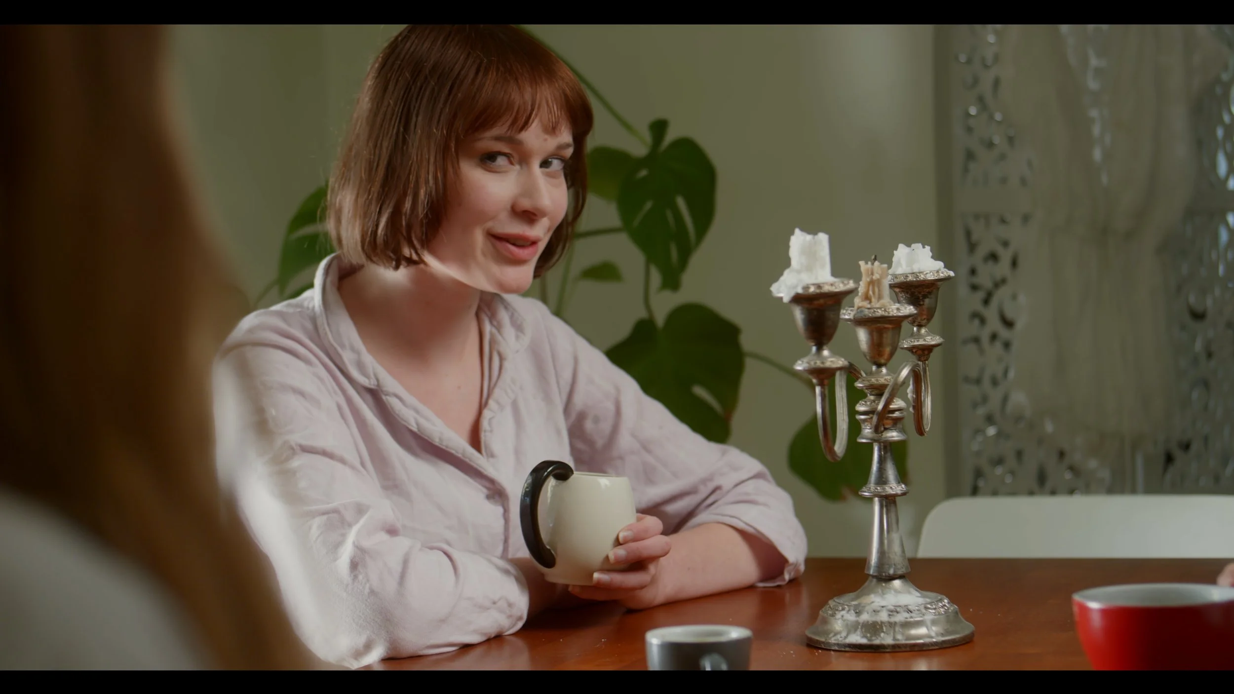 Woman sitting at a table with a white mug, next to a silver candelabra with melted candles; a potted plant and patterned divider in the background.