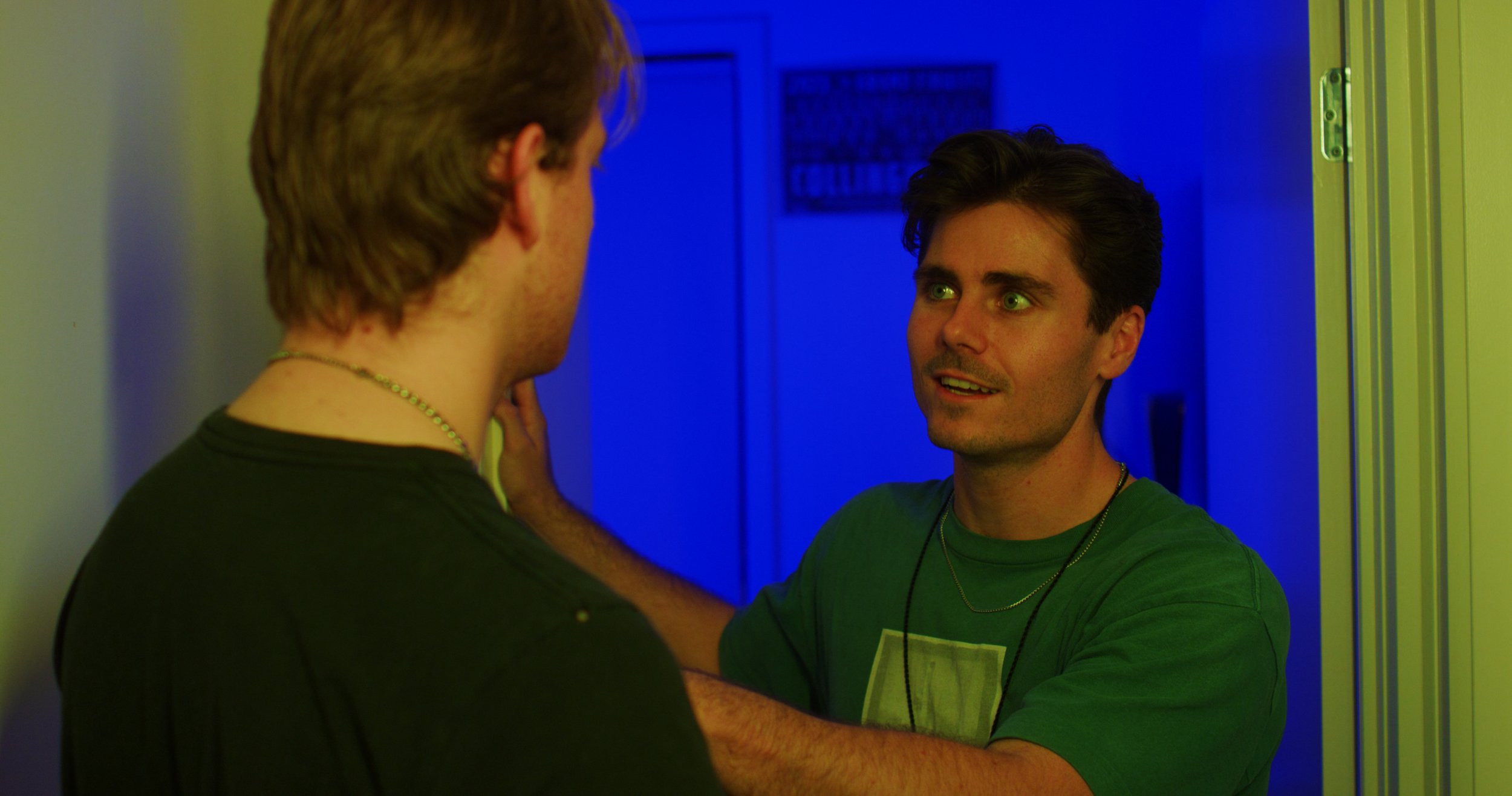 Two men in conversation in a dimly lit room with blue and green lighting.