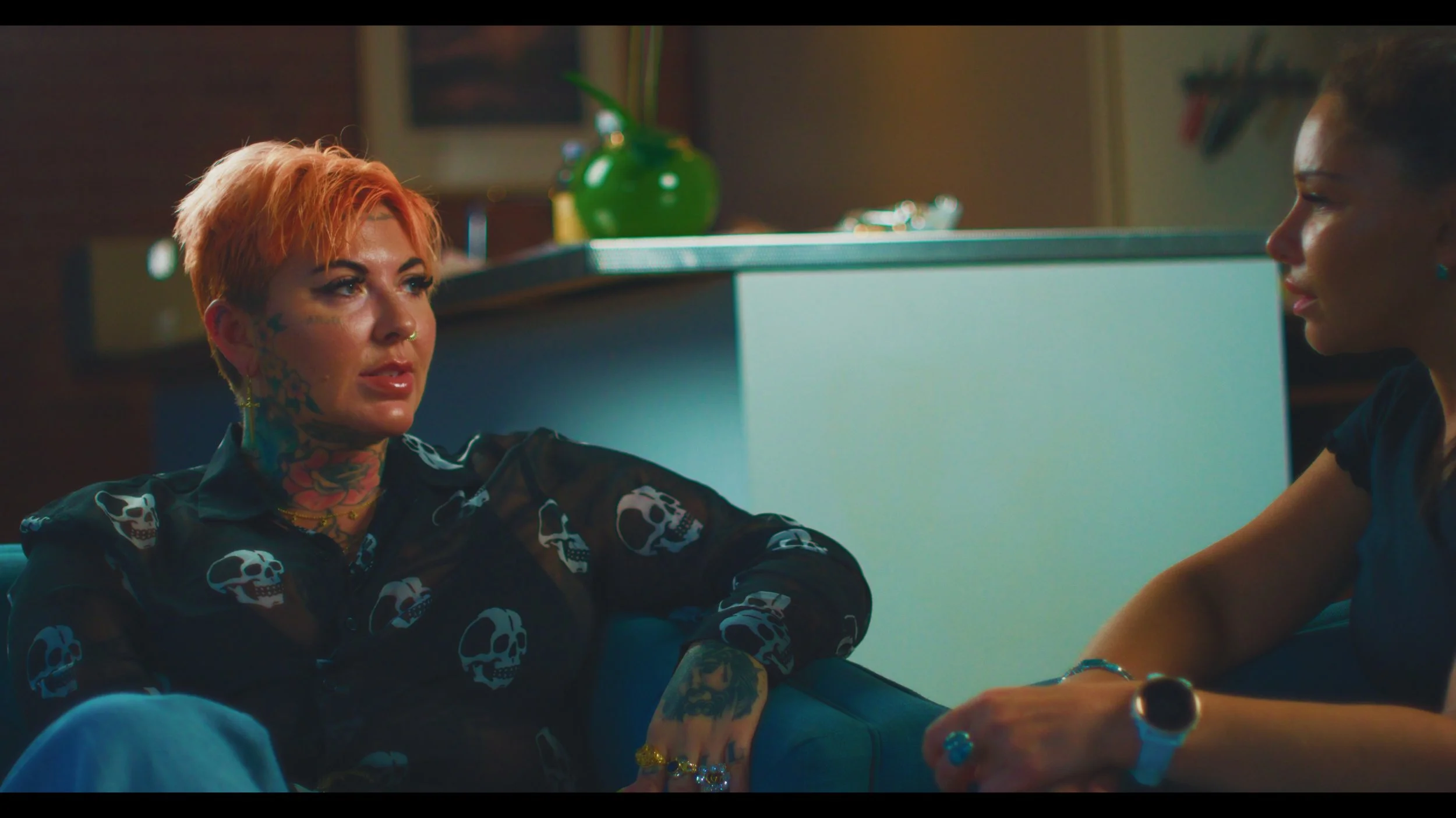 Two people sitting and talking in a living room, one with short orange hair, tattoos, and a sheer skull-patterned shirt, the other wearing a black shirt and a smartwatch.
Interview set up and lighting for doco.