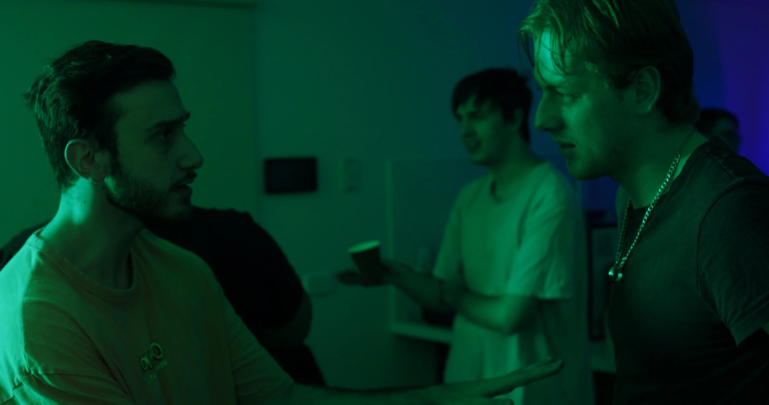Three men in a dimly lit room, one holding a cup, talking animatedly with green lighting.