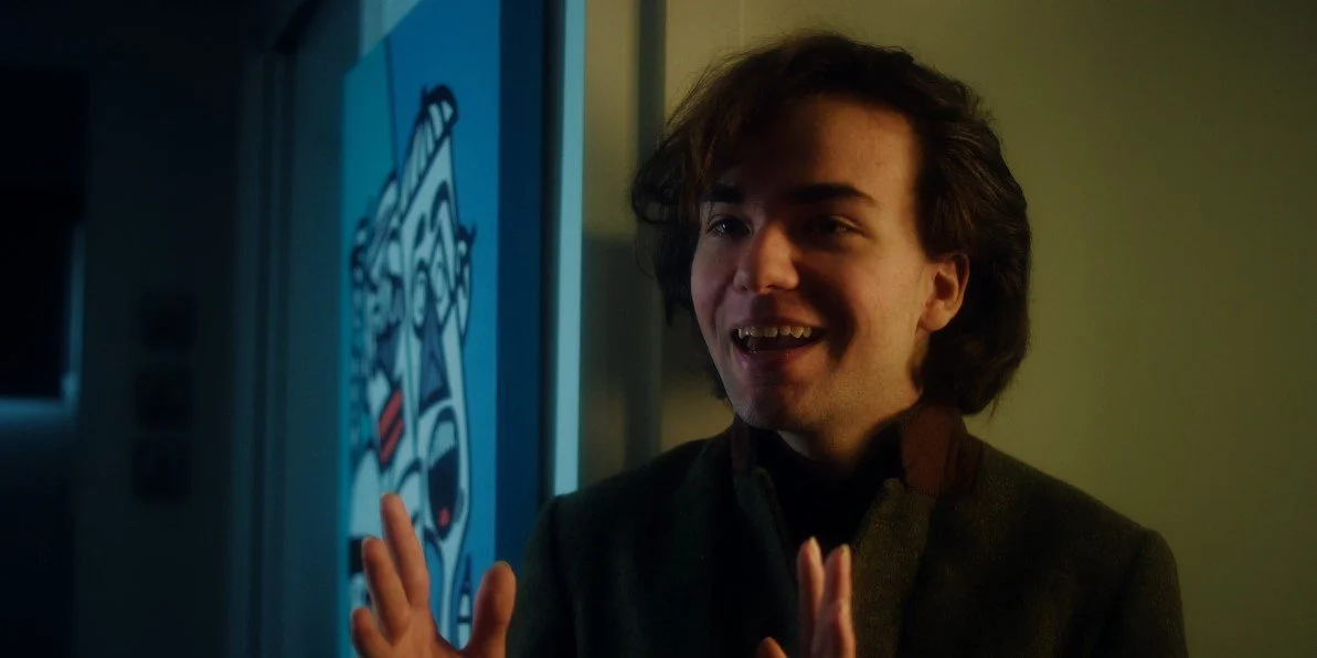 A person with medium-length hair smiling and gesturing with hands in front of a blue artwork featuring abstract faces.