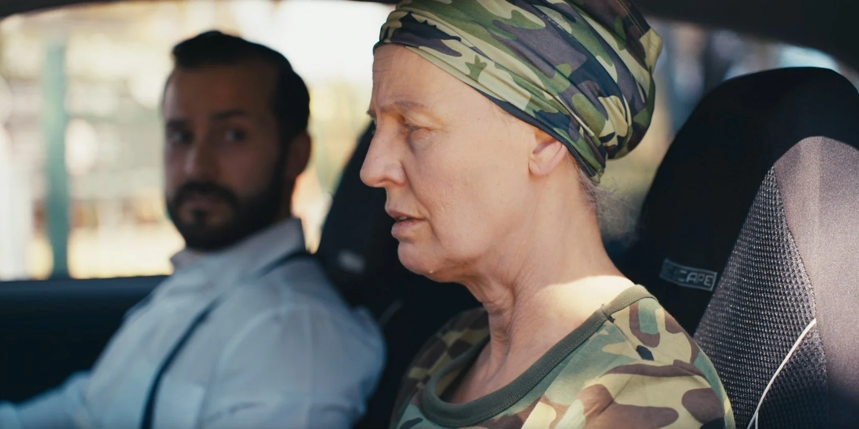 An elderly woman wearing a camouflage headscarf and shirt sitting in a car, with a man in the background seated next to her.