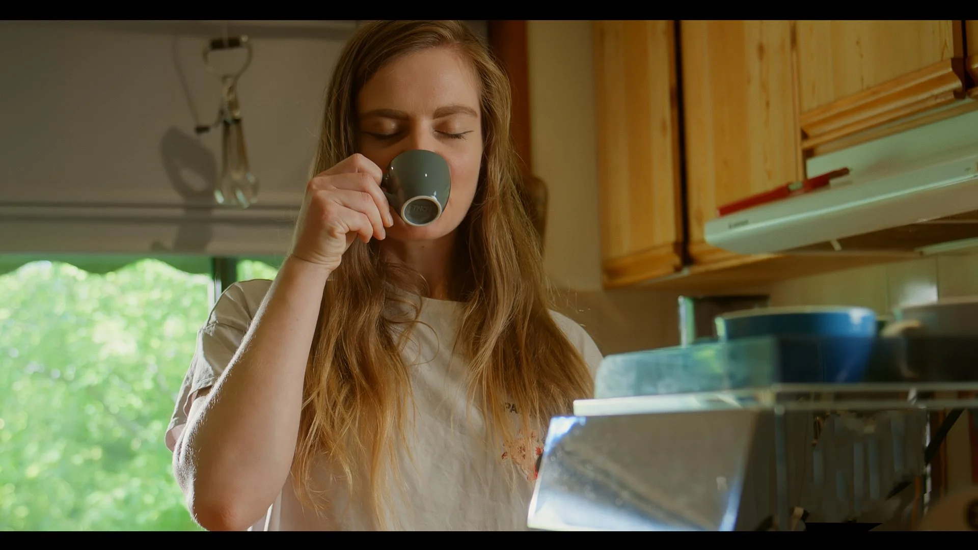Woman drinking coffee in a kitchen with sunlight through a window.