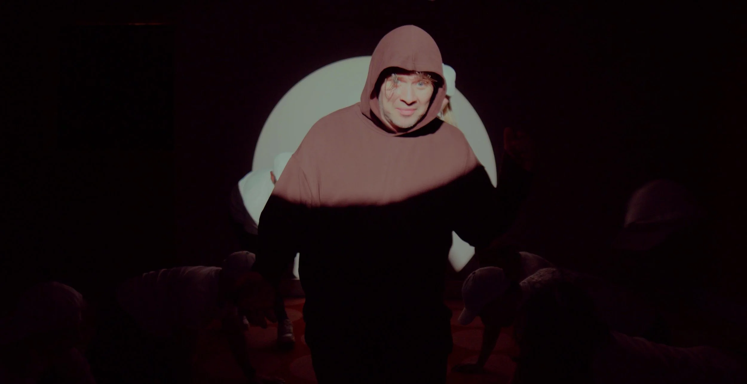 A person in a hooded sweatshirt partially illuminated in dim lighting with a bright circular background. Other people are in crouching positions wearing white clothing.
