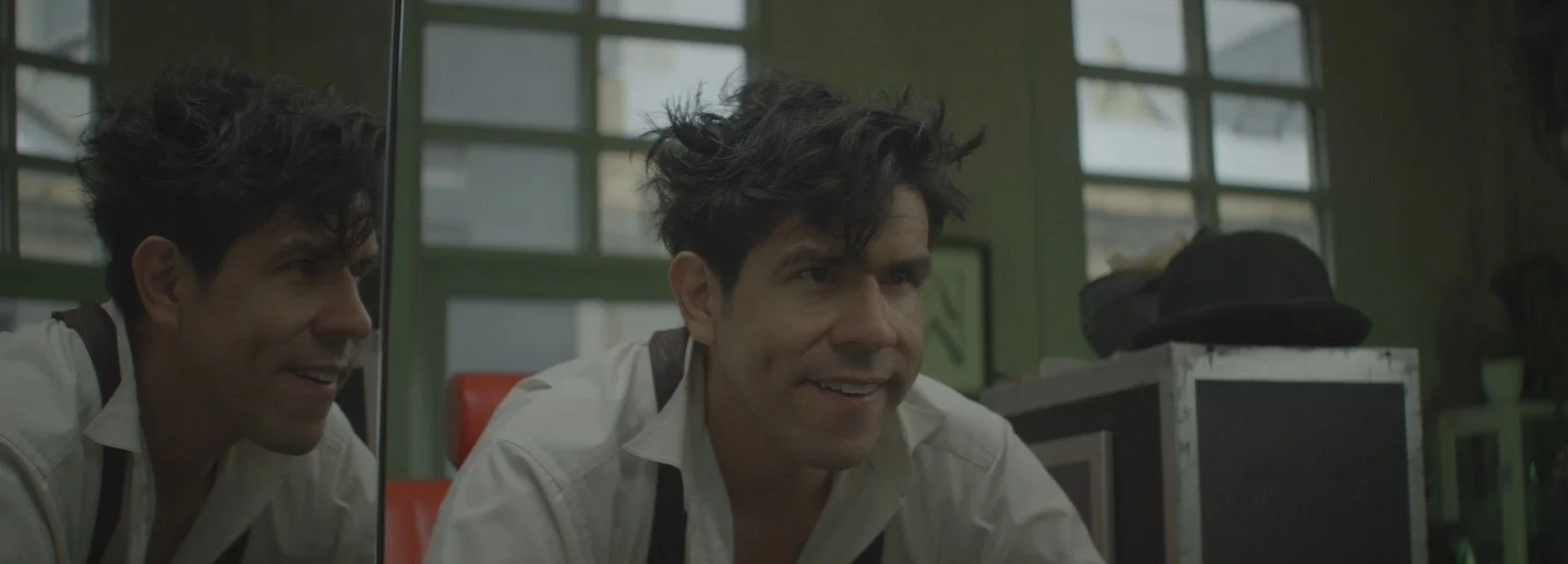 Man with tousled hair in white shirt and dark suspenders smiling in front of a mirror, reflecting his face, inside a room with large windows and a hat on a nearby object.
Music video lighting and cinematography