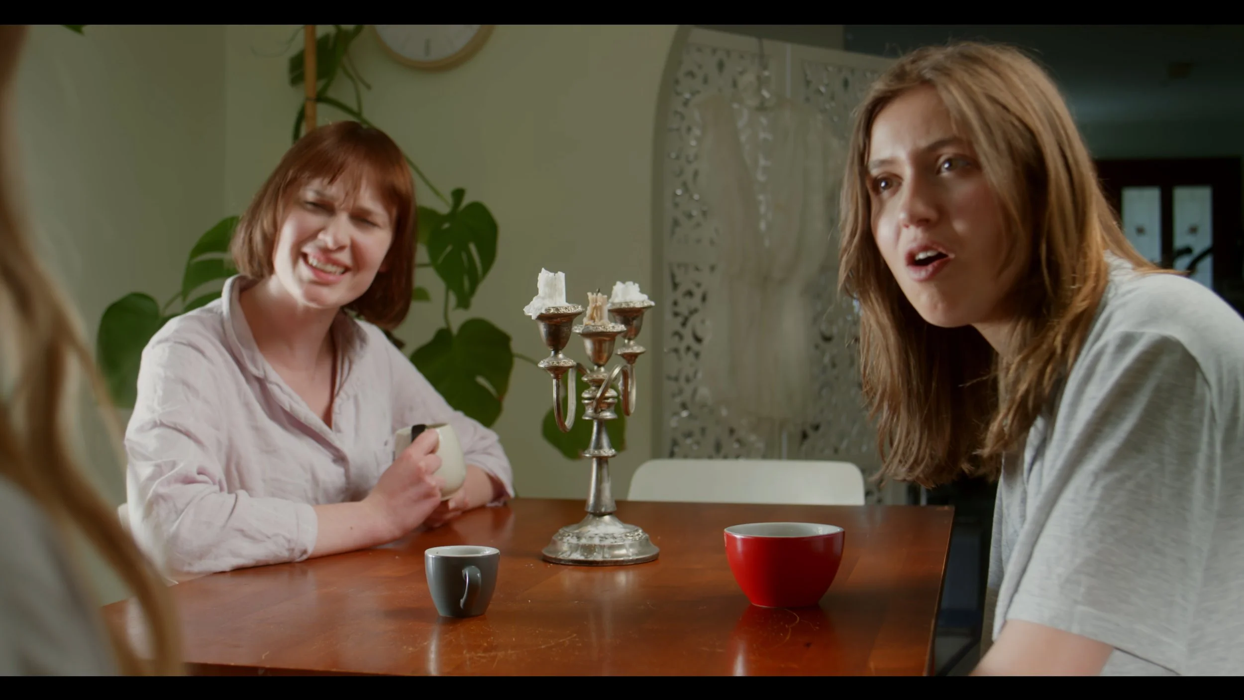 Two women sitting at a table with cups and a candelabra, engaged in conversation.