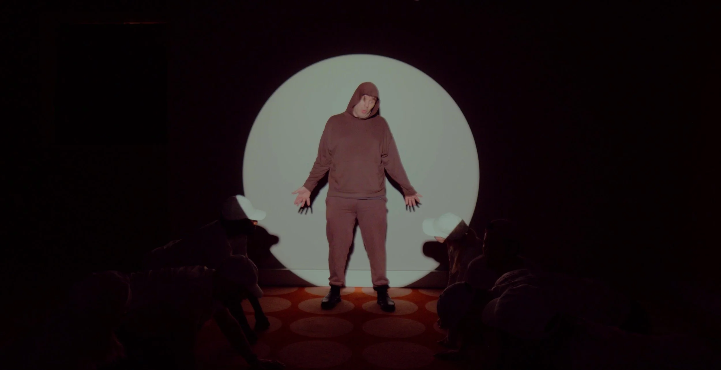 Person in hooded outfit standing in spotlight with others crouching around