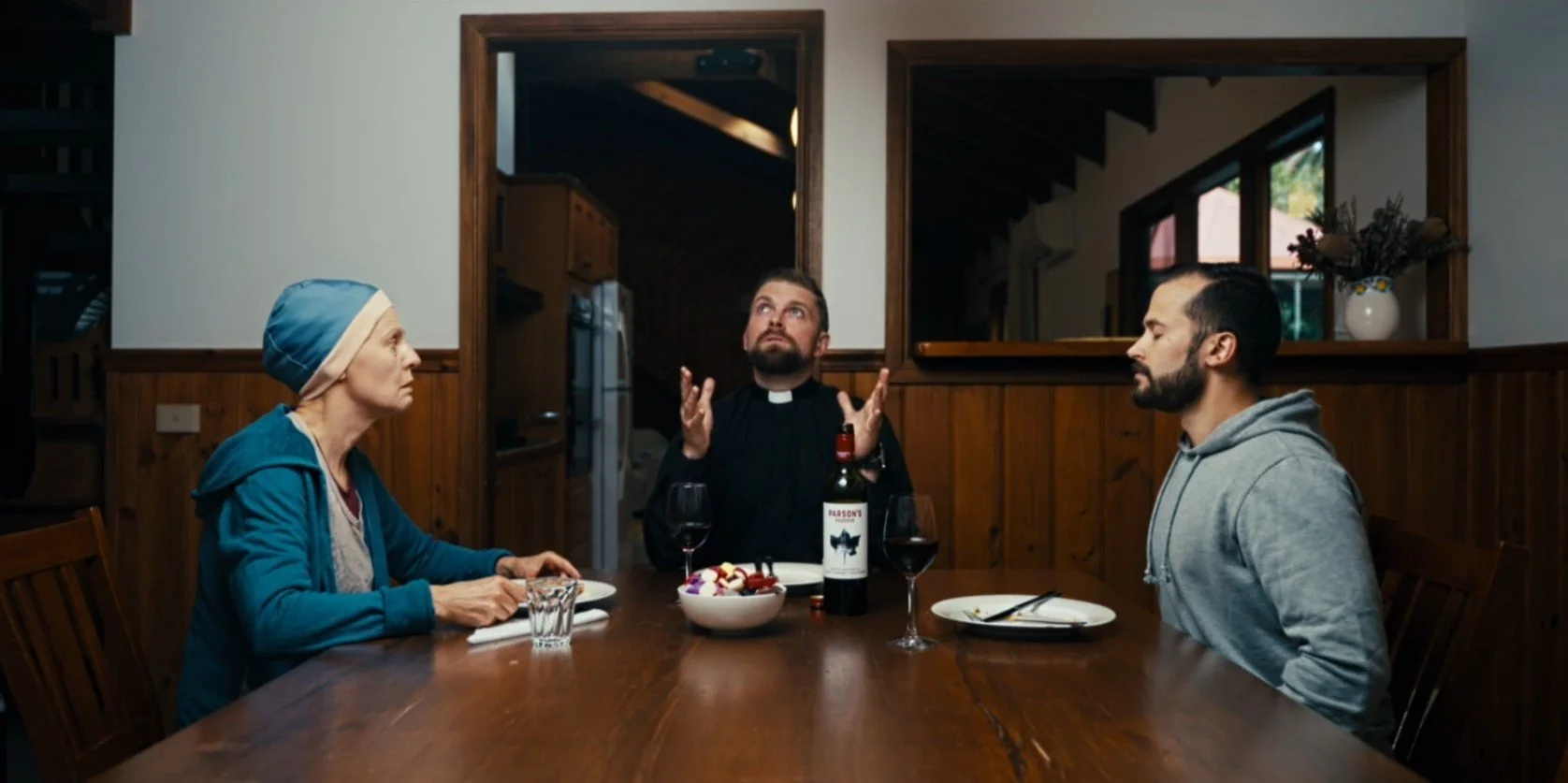 Three people sitting at a dining table, two men and a woman. One man is dressed as a priest, looking upward with hands raised. The woman wears a headscarf, and the other man is in a gray hoodie. A bottle of wine, glasses, and a bowl of salad are on the table.