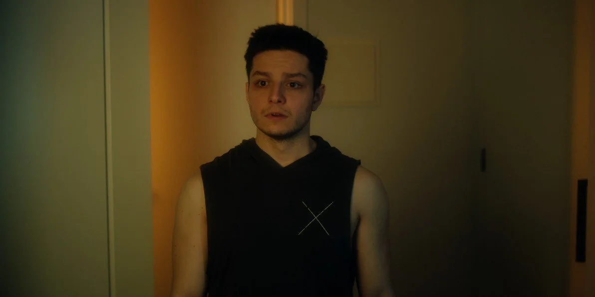 A person with short dark hair, wearing a sleeveless black top, standing in a dimly lit room.