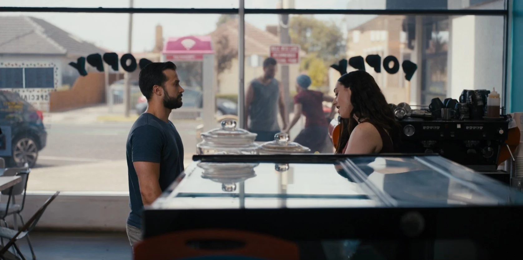 Two people talking at a cafe counter with two people outside the window.