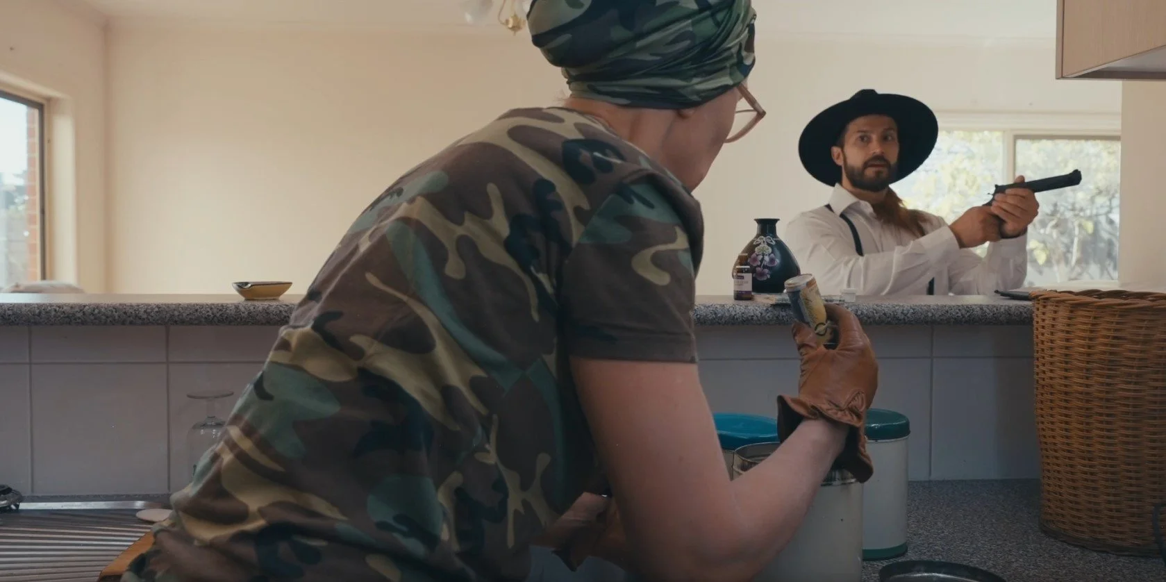 Person in camouflage outfit holding can in kitchen, person in background with black hat and gun.