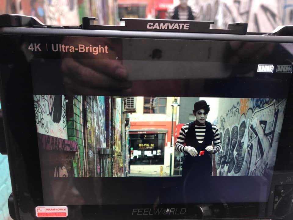 Camera monitor displaying video footage of a street scene with graffiti and a mime wearing a striped shirt and hat.