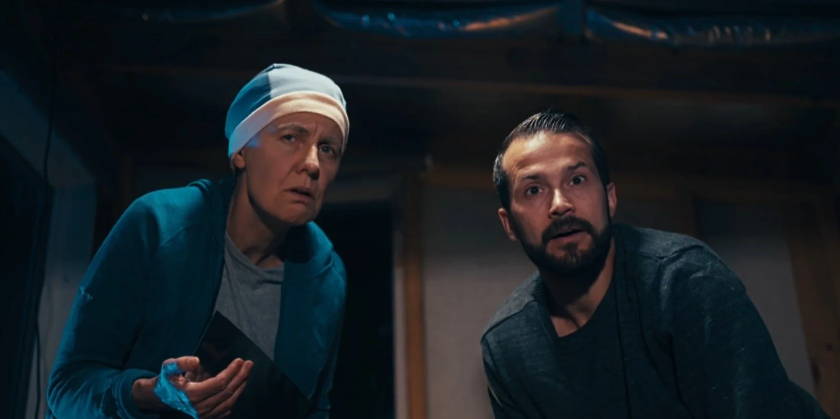 Two people indoors, one wearing a blue jacket and a headscarf, the other with a beard and gray shirt, both looking surprised.