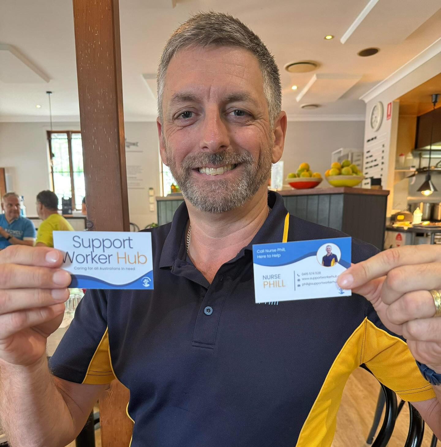 A man with short grey hair and a short grey beard in a navy and gold polo holding up two business cards.