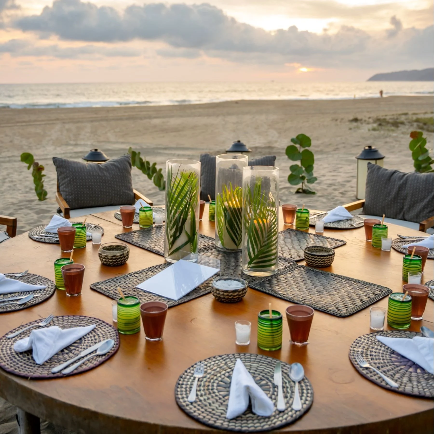Outdoor dining setup on a beach with a circular wooden table, wicker placemats, white cloth napkins, glasses, and palm leaves in vases. Evening sunset and ocean in the background.