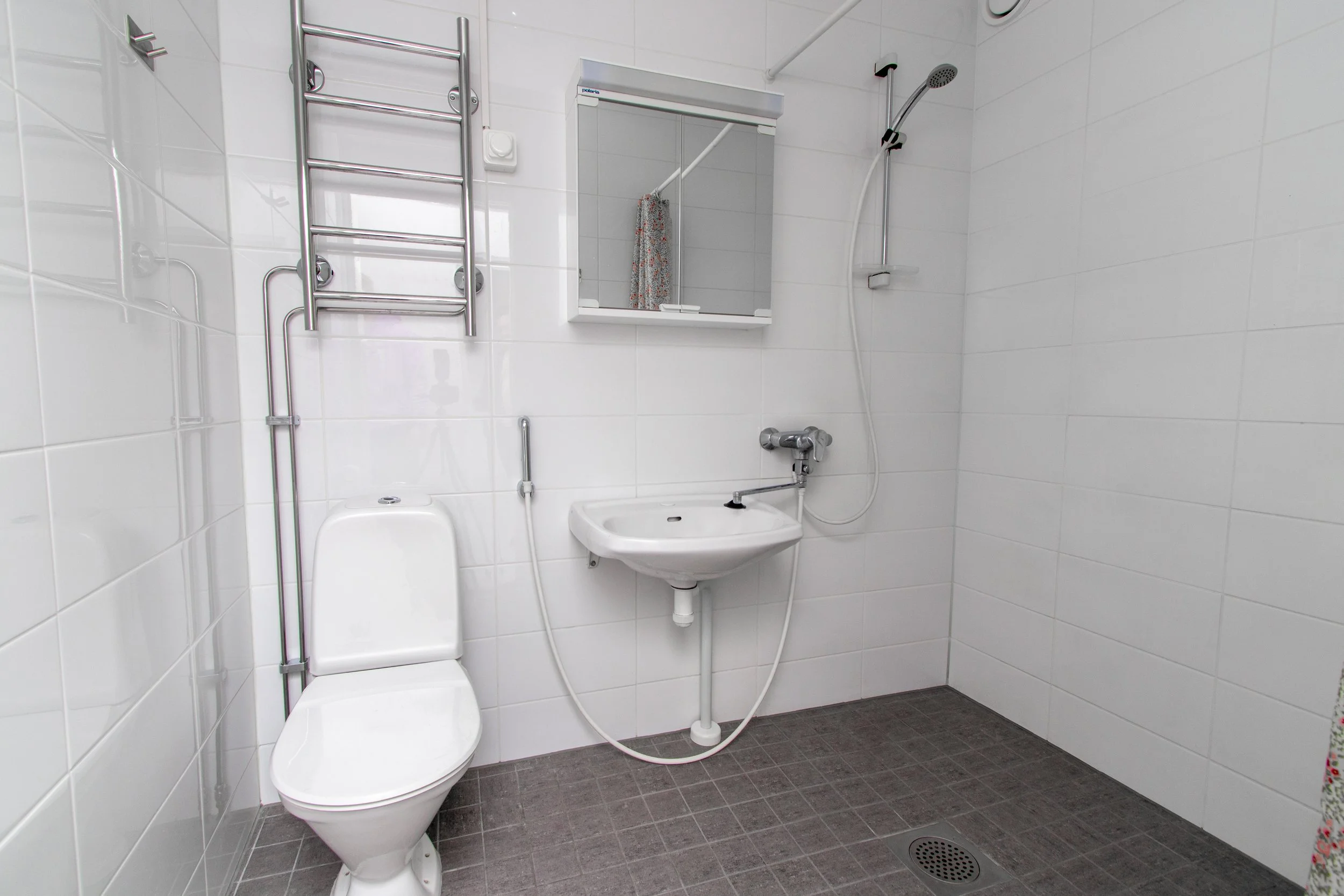 Pieni kylpyhuone, jossa on wc, pieni lavuaari, peili ja käsisuihku, valkoiset laatoitetut seinät ja tummat lattialaatat.