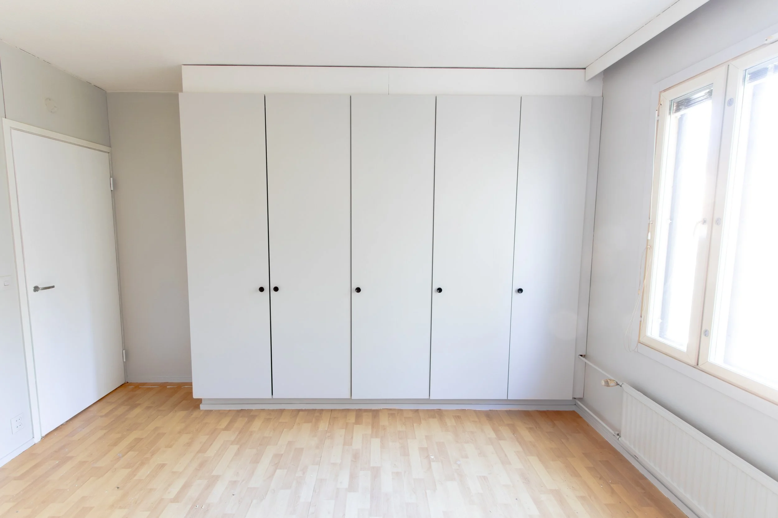 Tyhjä huone, vaaleat seinät, suuri valkoinen kaapisto, parkettilattia, ikkuna ja raita pattereiden kanssa seinässä.