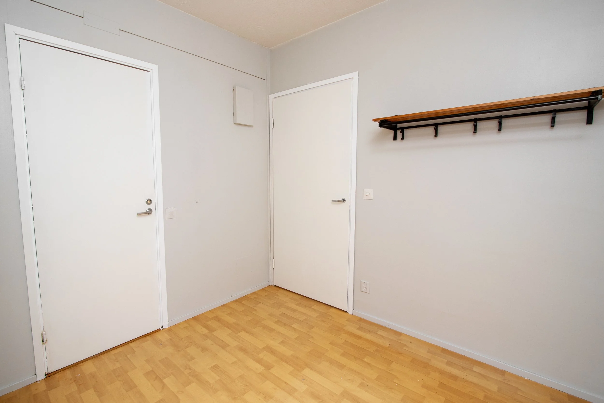 Tyhjä huone, valkoiset seinät ja kaksi ovea, yksi vasemmalla ja yksi oikealla, ja puiset lattialankut. Yhdellä seinällä on seinähylly ilman tavaroita.