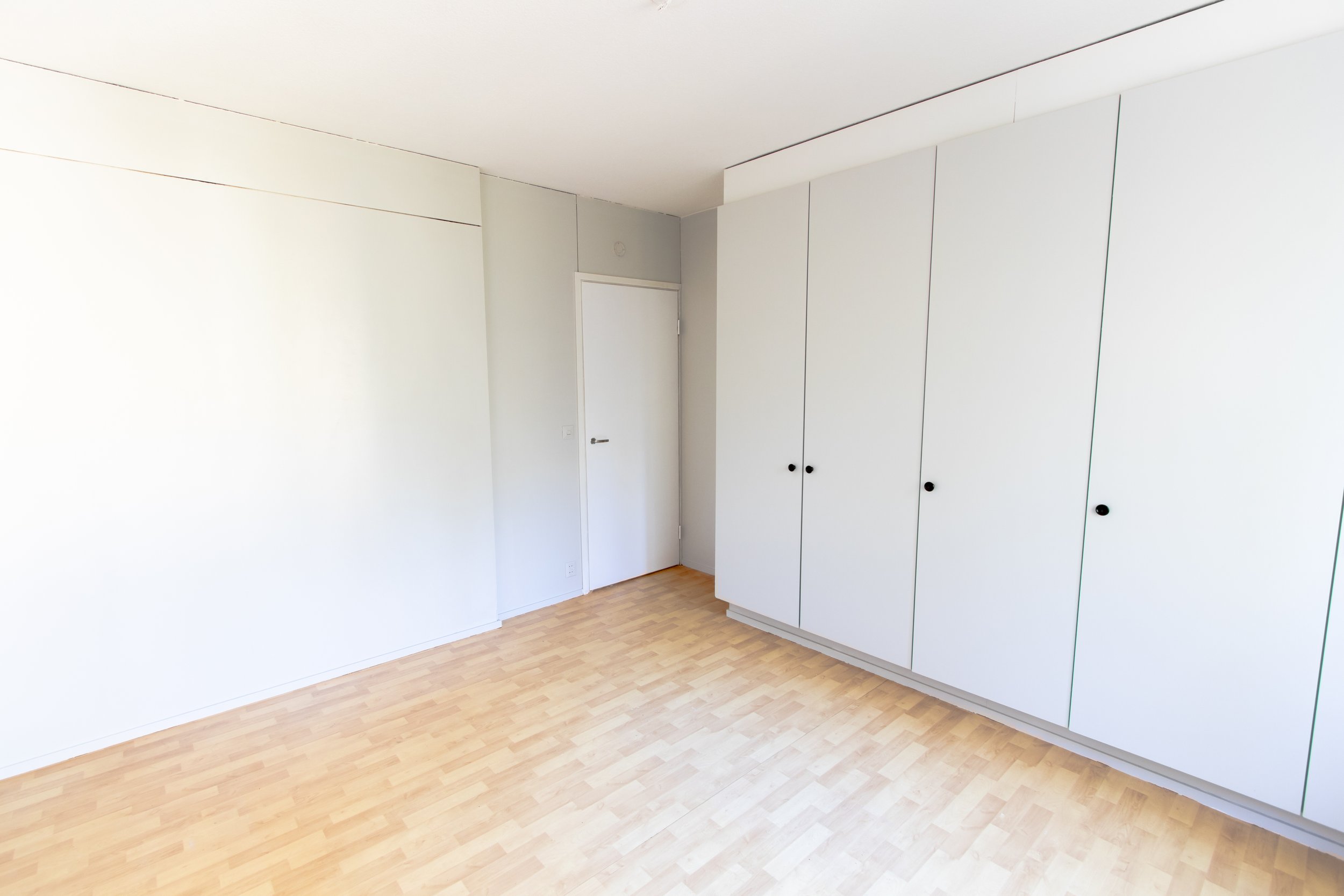 Tyhjä huone, valkoinen seinä, säilytystankki ja ovi, laminaattilattia.