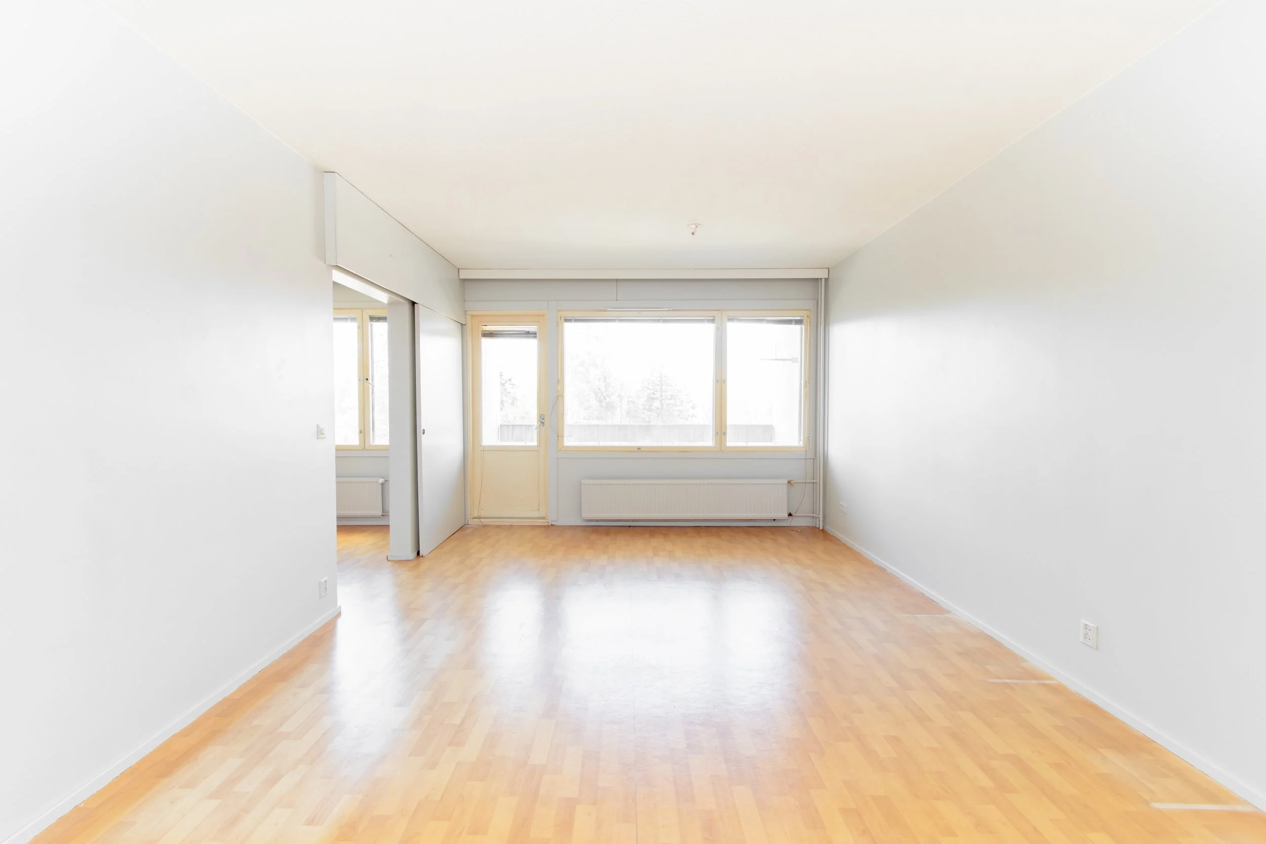 Avaamaton huone, jossa puulattia ja suuret ikkunat, valkoinen seinät ja ovi johtamassa toiseen huoneeseen.