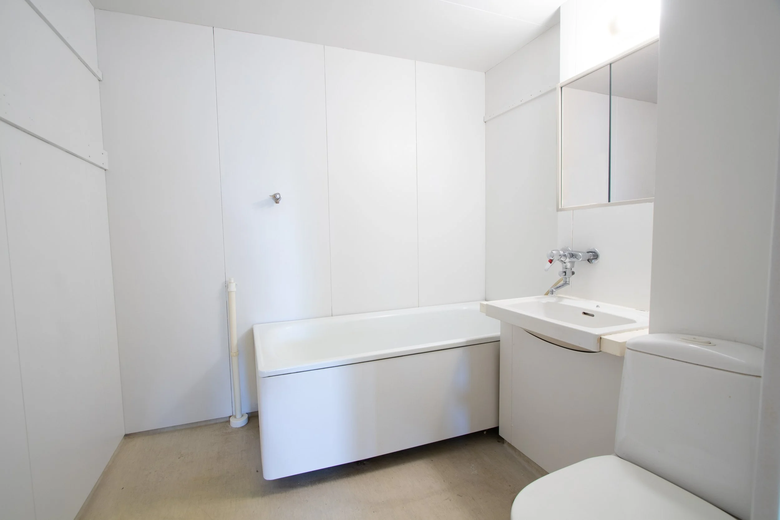 Valkoinen kylpyhuone, jossa kylpyamme, pesuallas ja WC-istuin, seinät ja huonekaluja, pieni peilejä.