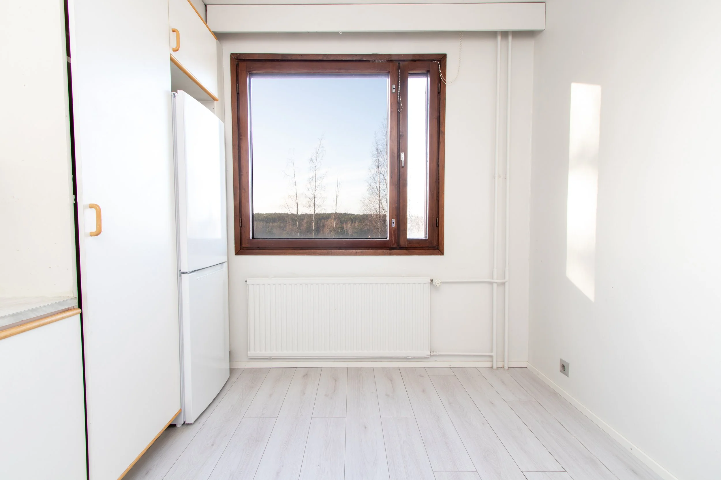 Tyhjä keittiön ikkuna, valkoinen seinä ja laminaattilattia.