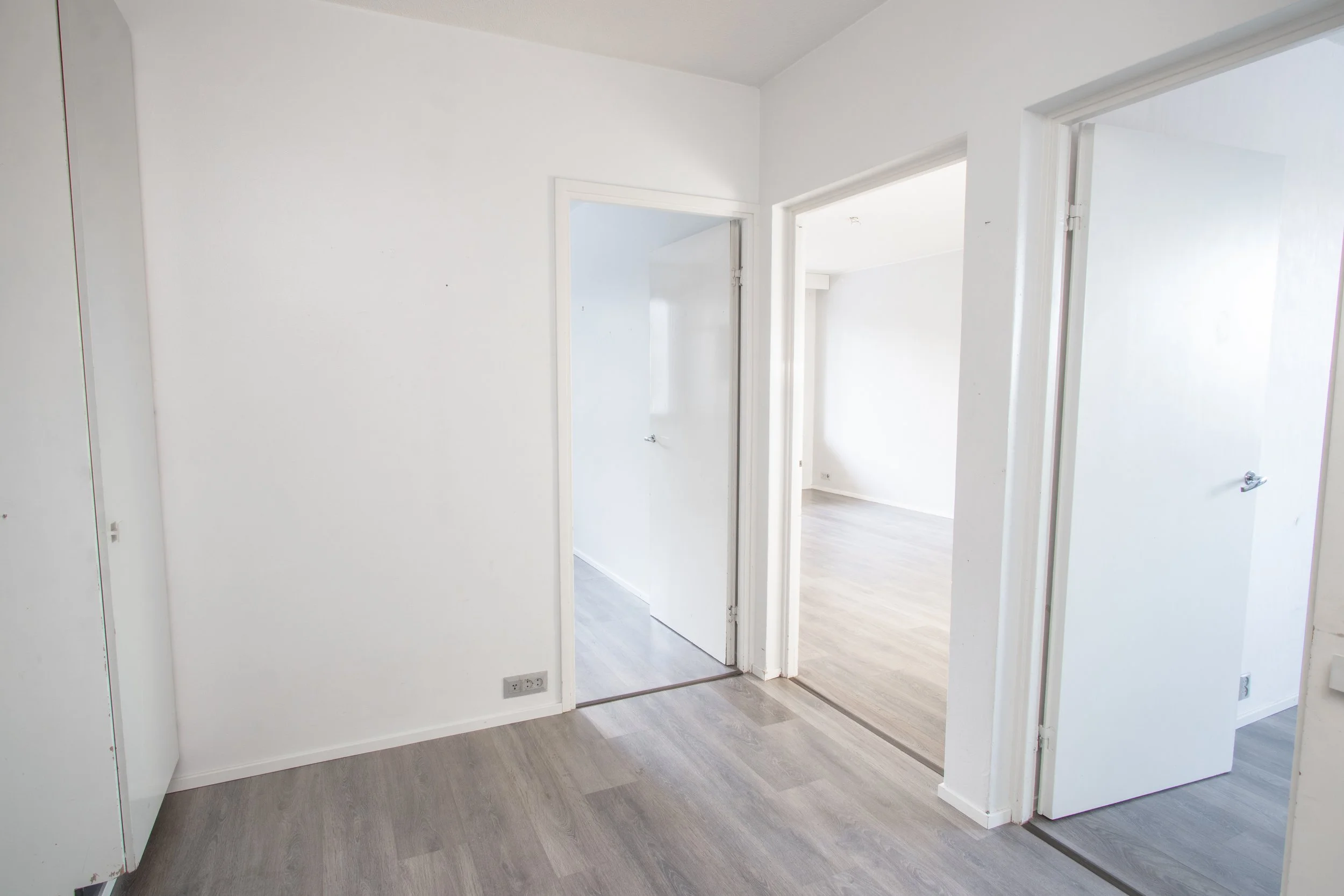 Valkoinen huone, jossa kolme ovea johtaa muihin huoneisiin, ja harmaat laminaattilattiat.