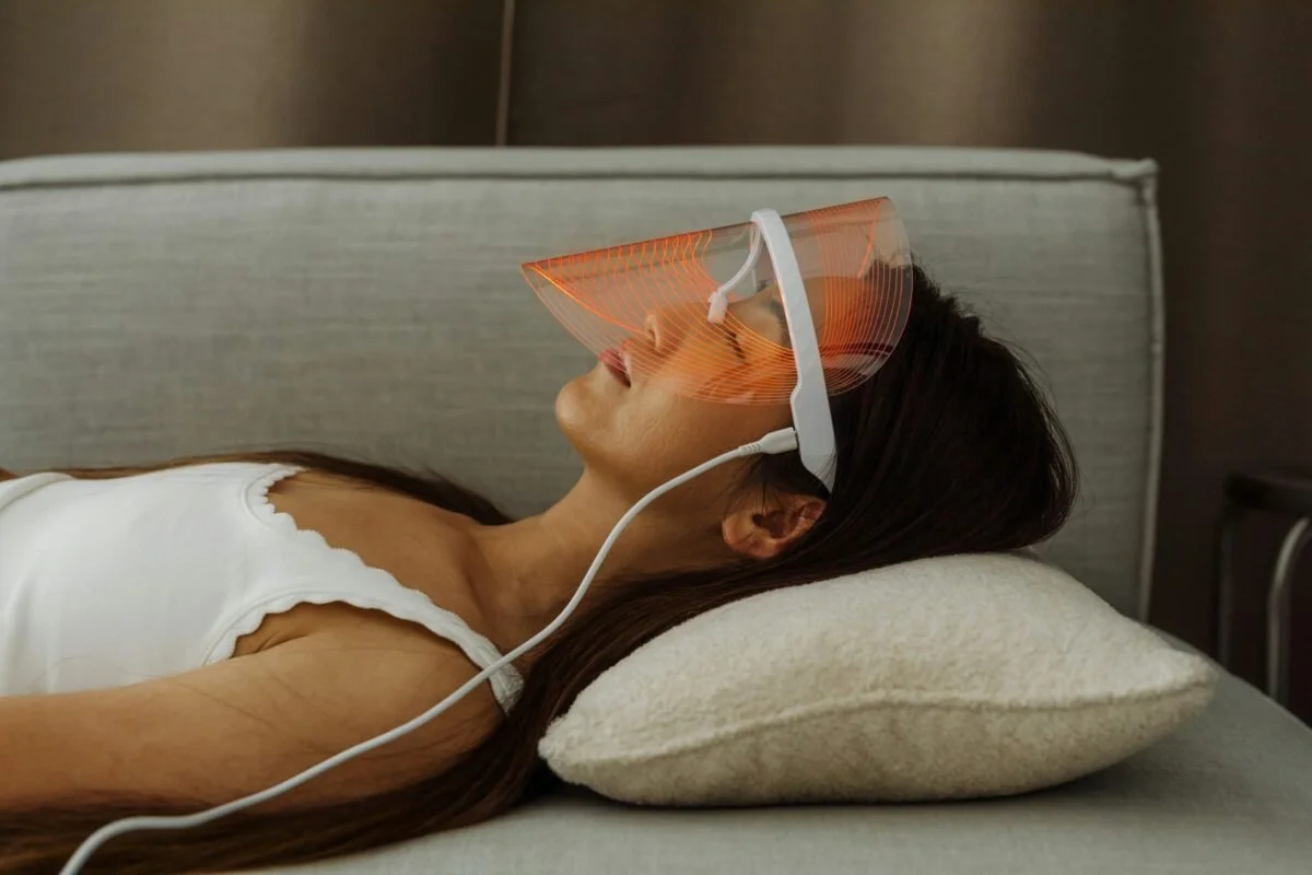 Femme allongée sur un lit portant un masque de luminothérapie LED rouge et orange.