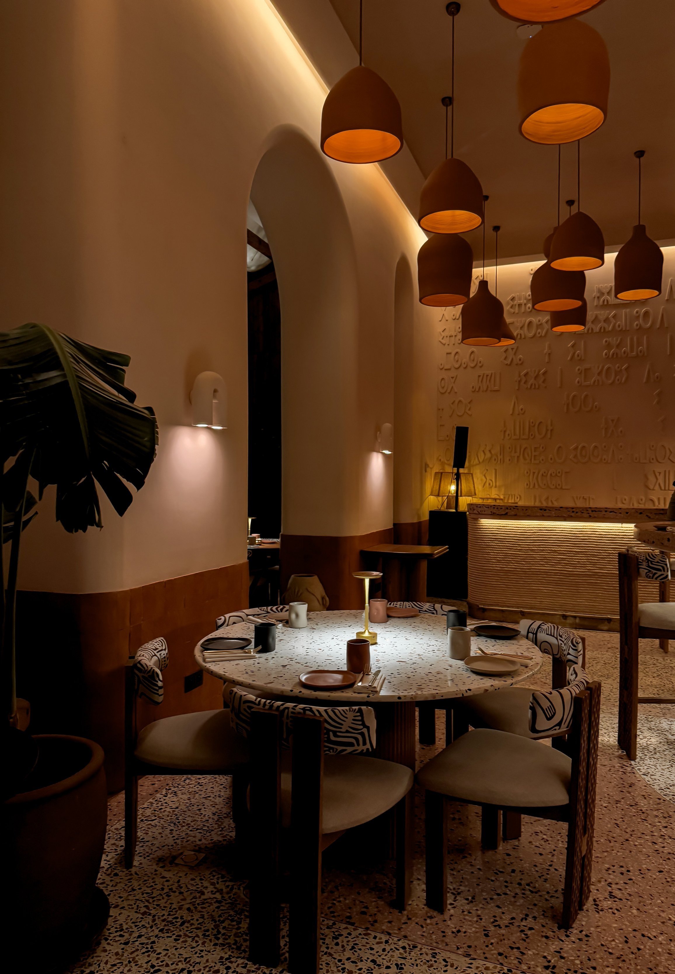 Une salle de restaurant élégante avec un éclairage tamisé, un mur décoré avec des lettres en relief, une table ronde avec des assiettes, des verres et des bougeoirs. Des lampes suspendues au plafond ajoutent une ambiance chaleureuse.