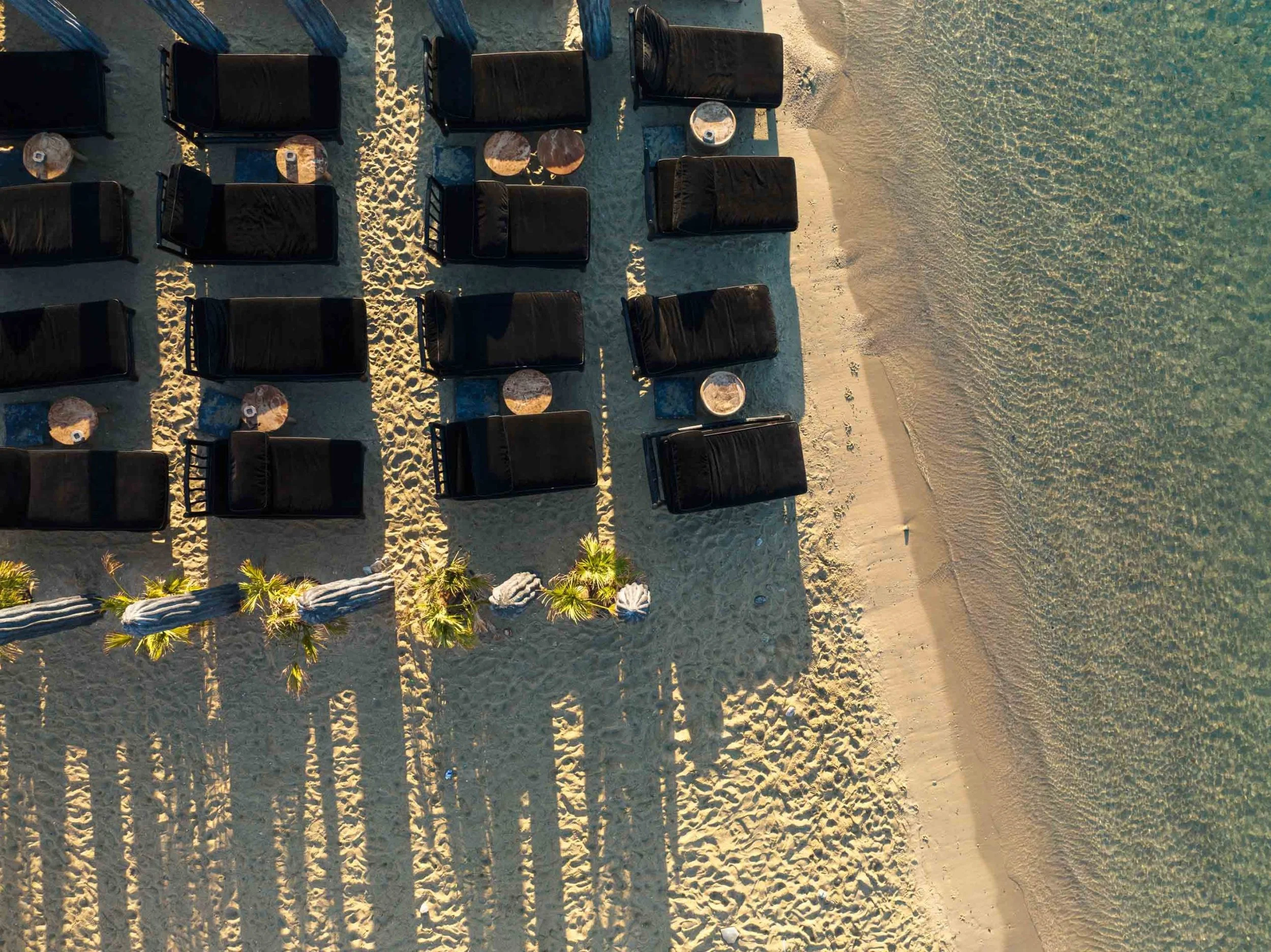 Chaises longues noires avec petites tables ronde en marbre sur une plage de sable fin, vue aérienne au coucher du soleil.