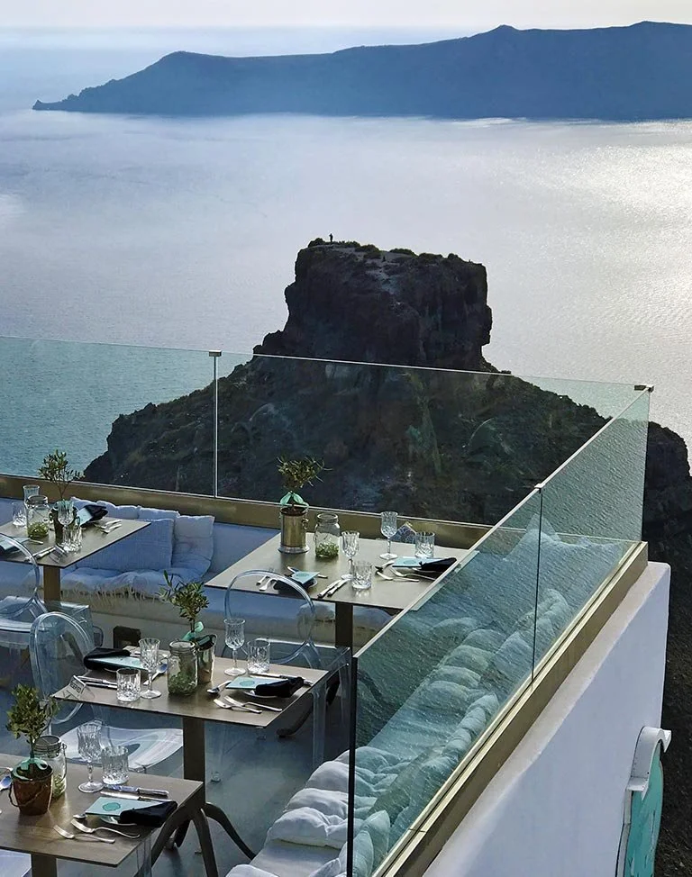 Vue d'une terrasse avec tables et chaises, donnant sur une mer calme et une falaise rocheuse avec un promontoire, dans un paysage de côte méditerranéenne.