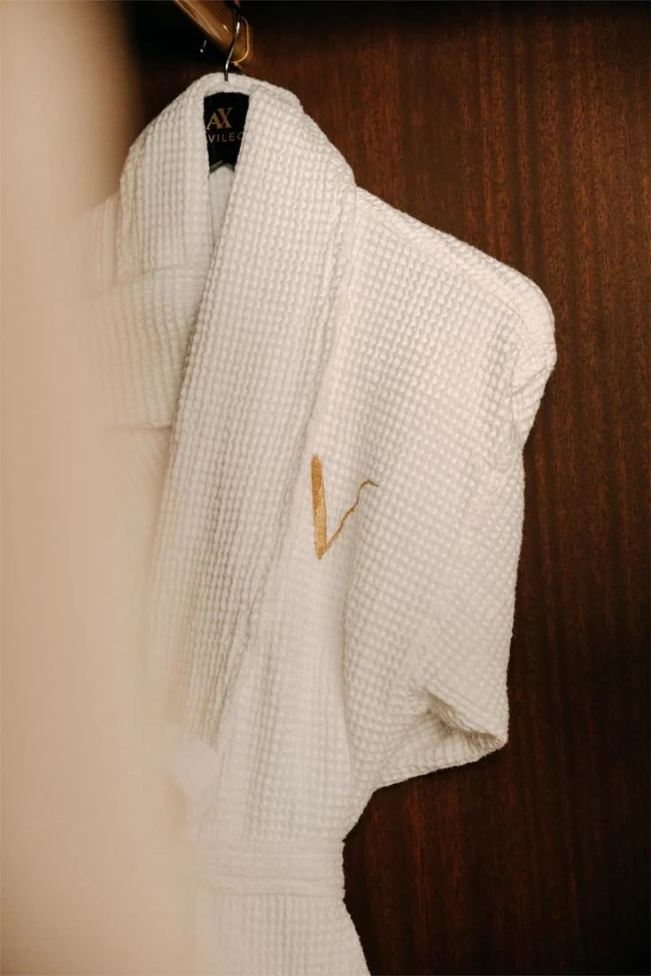 Peignoir blanc texturé suspendu à une patère en bois, avec logo brodé doré sur la poitrine.