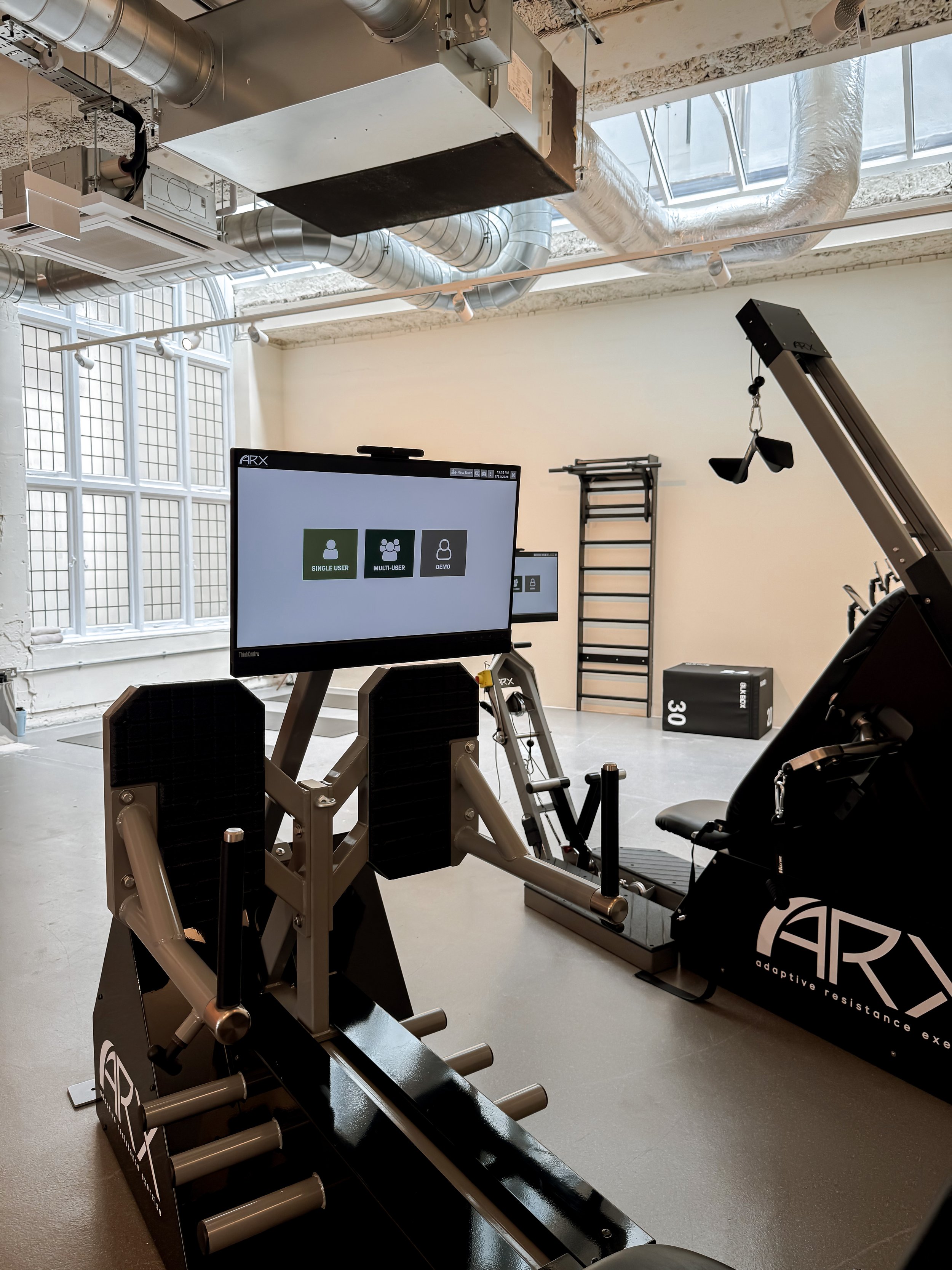Équipement d'entraînement en salle de sport avec un vélo elliptique, un panneau d'écran et des appareils de musculation, dans une pièce bien éclairée avec de grandes fenêtres et des conduits d'air visibles au plafond.