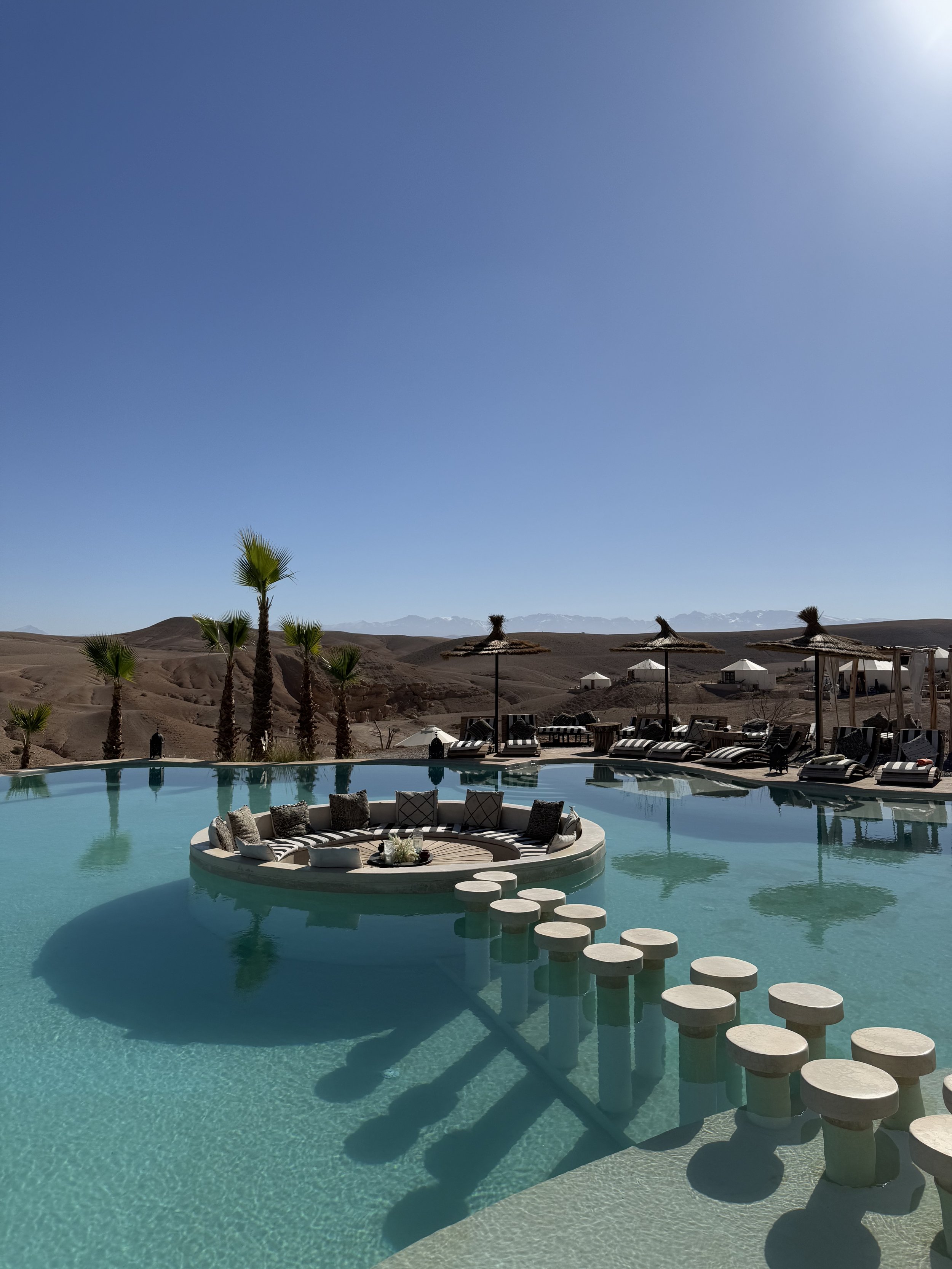 Piscine dans un désert avec des parasols en paille, des fauteuils et un paysage montagneux en arrière-plan, sous un ciel dégagé.