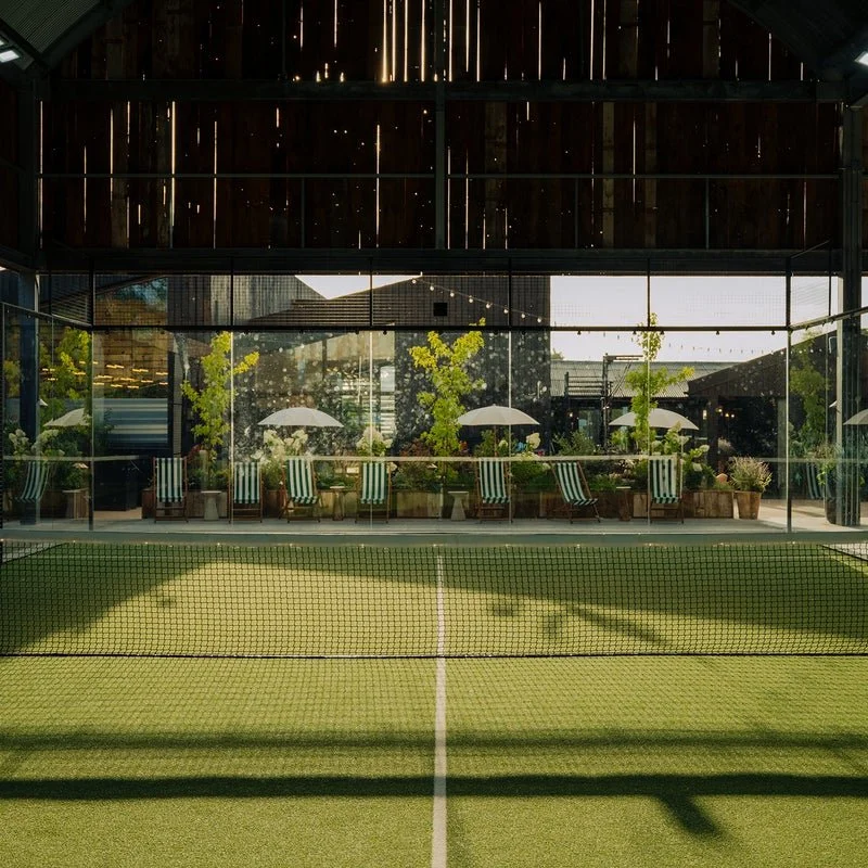 Un court de tennis en intérieur avec un filet au centre, entouré de fenêtres en verre, des plantes et des chaises avec parasols en fond.