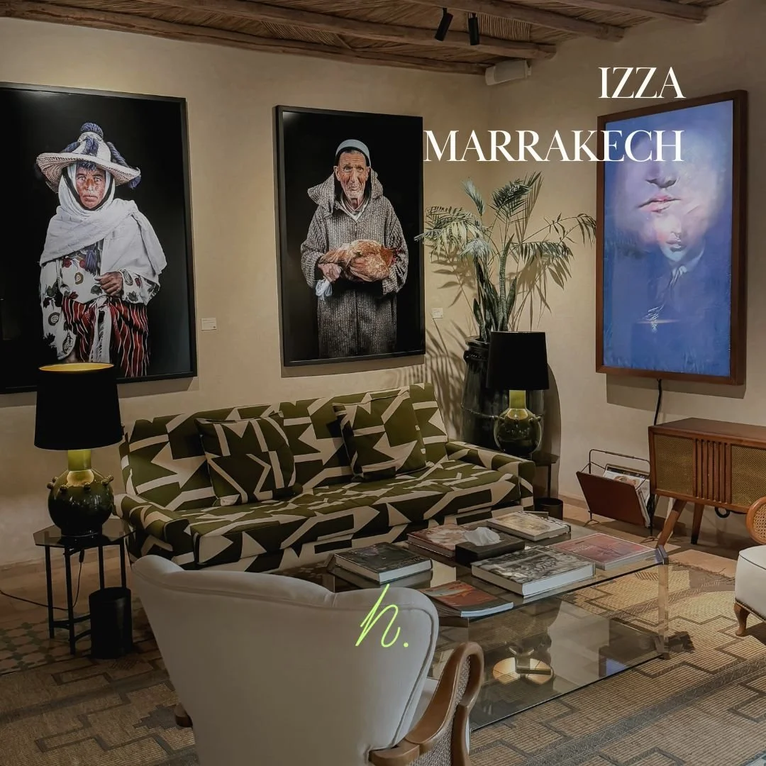 Une salle de séjour avec un canapé à motifs, deux lampes de table, une table basse en verre avec des livres, une plante verte, et des portraits encadrés accrochés au mur.