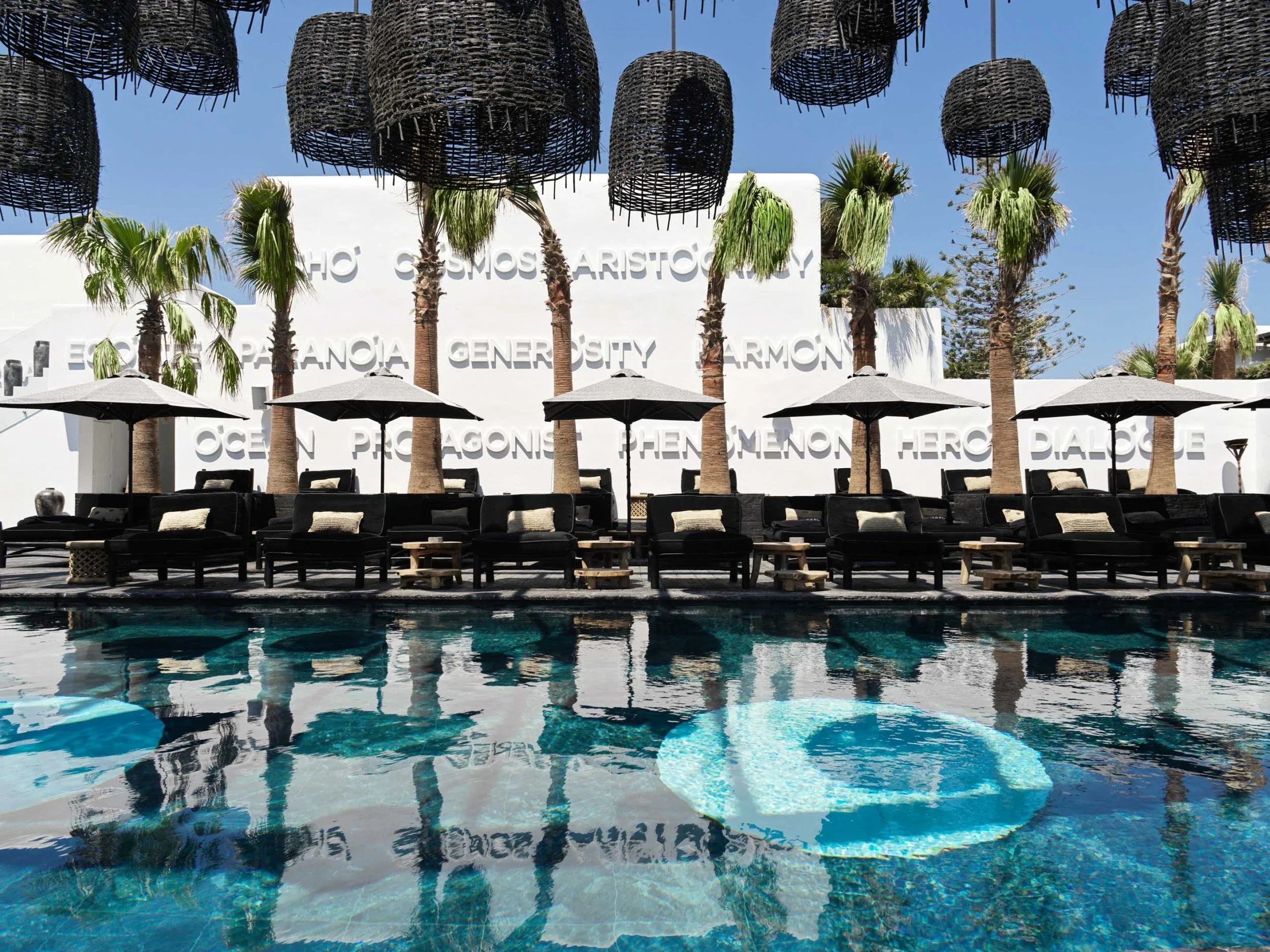Une piscine avec des chaises longues noires et des parapluies en verre gris en premier plan, des palmiers derrière, et un bâtiment blanc avec des écritures en relief, dont on voit quelques mots comme 'HERO', 'GODS', et 'HARMONY'.