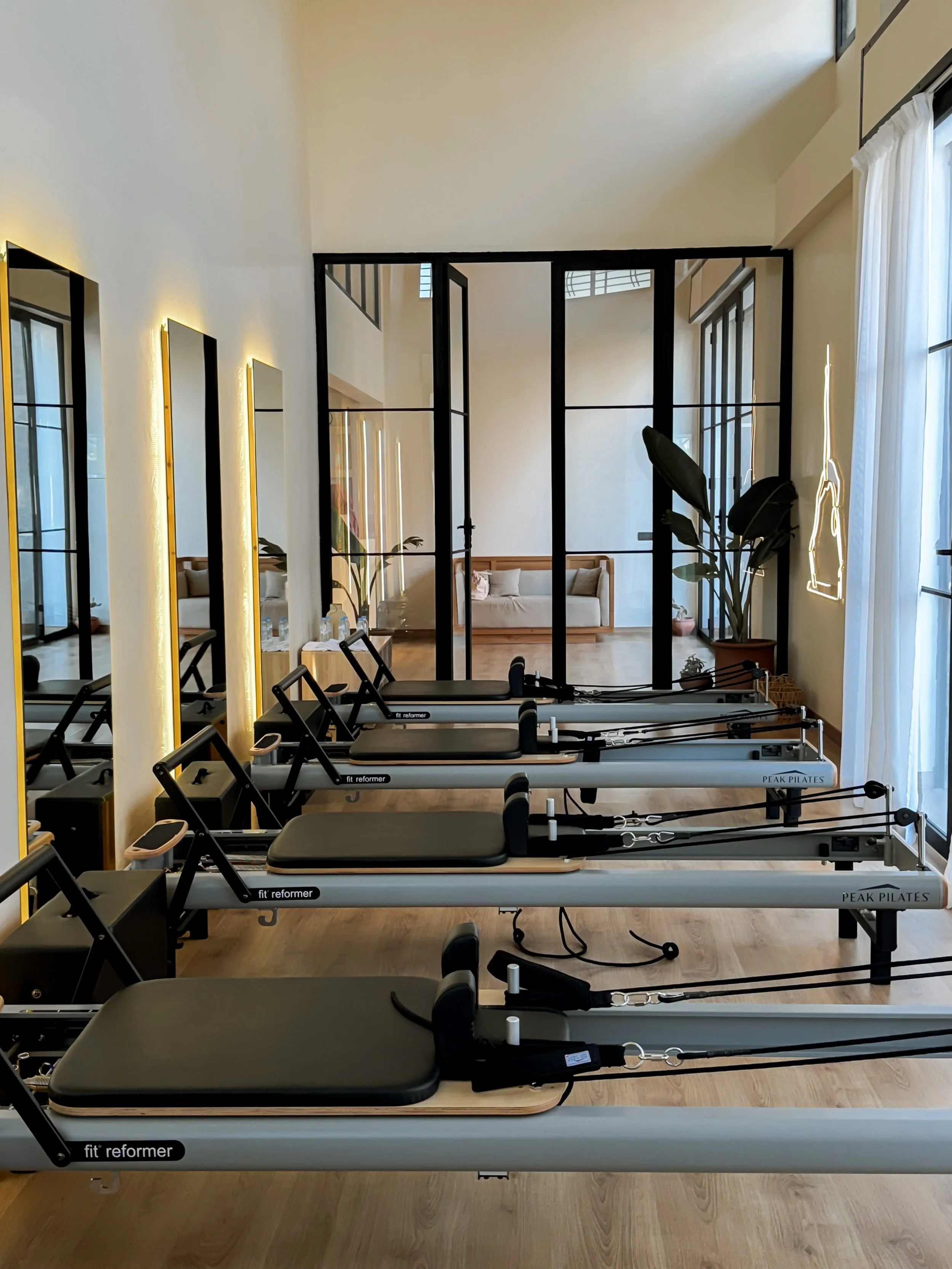 Salle de sport avec plusieurs reformers de Pilates alignés, miroirs sur le mur gauche, et un canapé en arrière-plan derrière une cloison en verre.