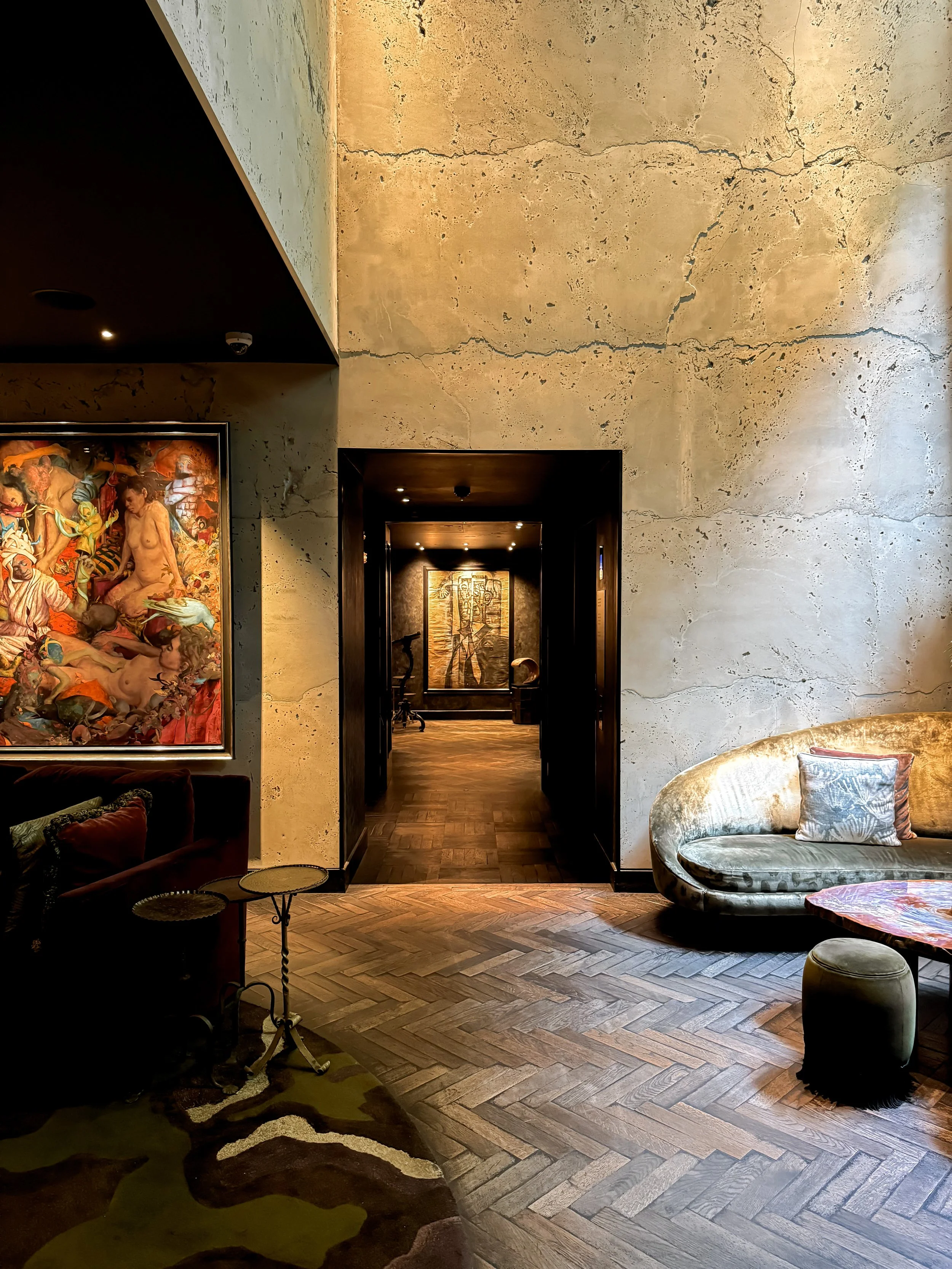 Intérieur d'un salon élégant avec un mur en pierre beige, une peinture colorée à gauche, un canapé en velours or et un fauteuil avec un coussin, un tapis en peau de vache, une petite table noire, et un couloir visible à travers une porte noire.