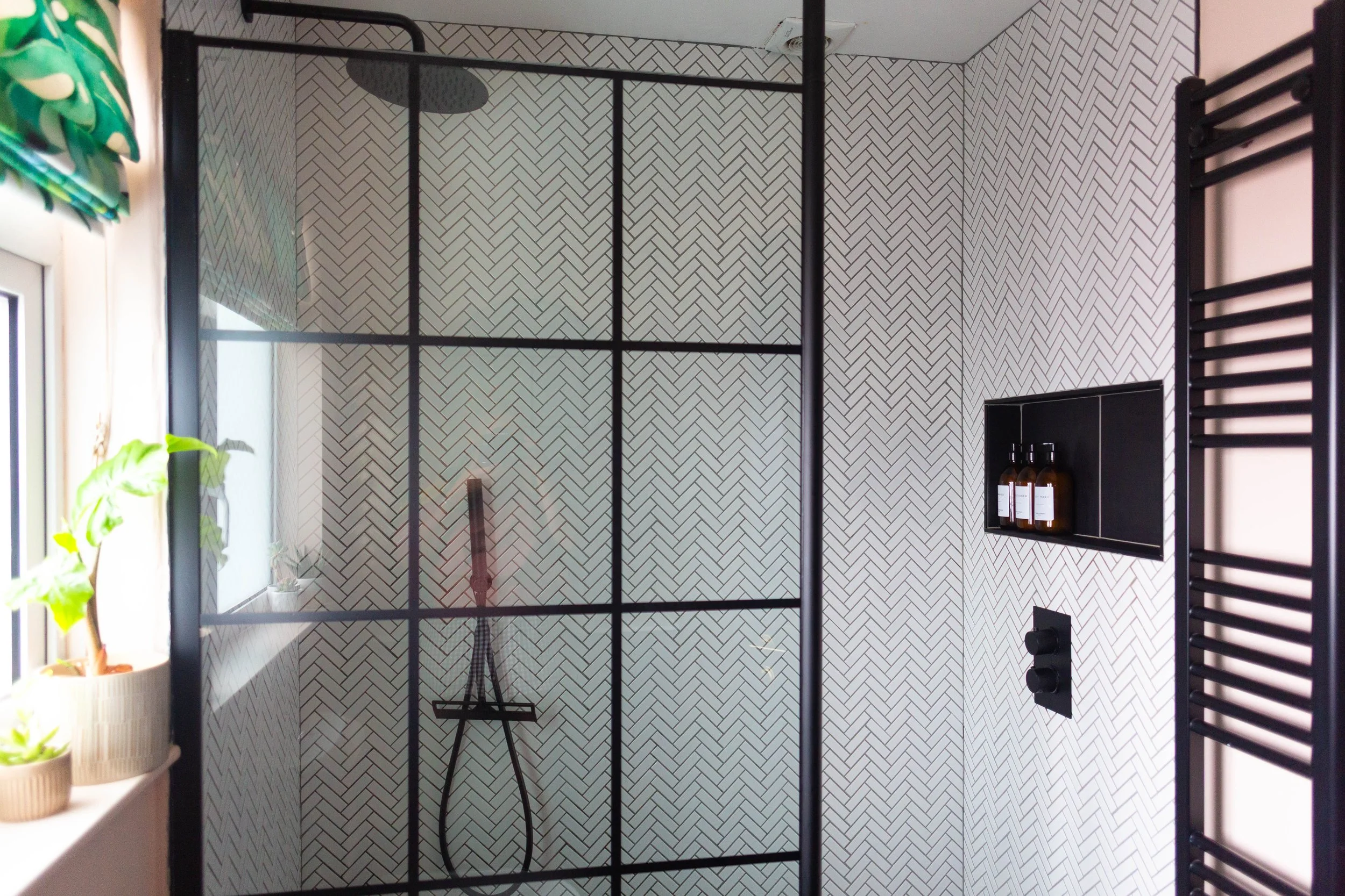 A modern bathroom with a walk-in shower featuring a black metal frame and a herringbone patterned tile wall. Inside the shower, there's a round rainfall showerhead and a black handheld shower. On the right wall, there's a black rectangular niche hold